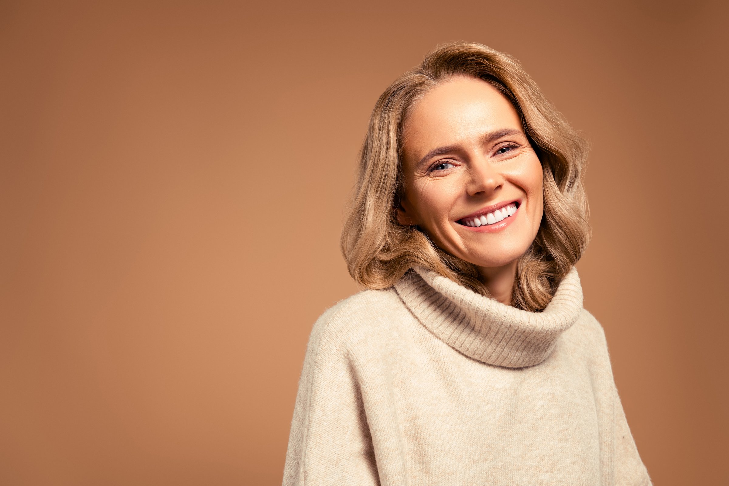 Mature blonde woman smiling warmly, wearing a stylish beige sweater, set against a soft beige backdrop, embodying elegance, sophistication, and an aura of positivity and beauty, showcasing natural charm and effortless grace.