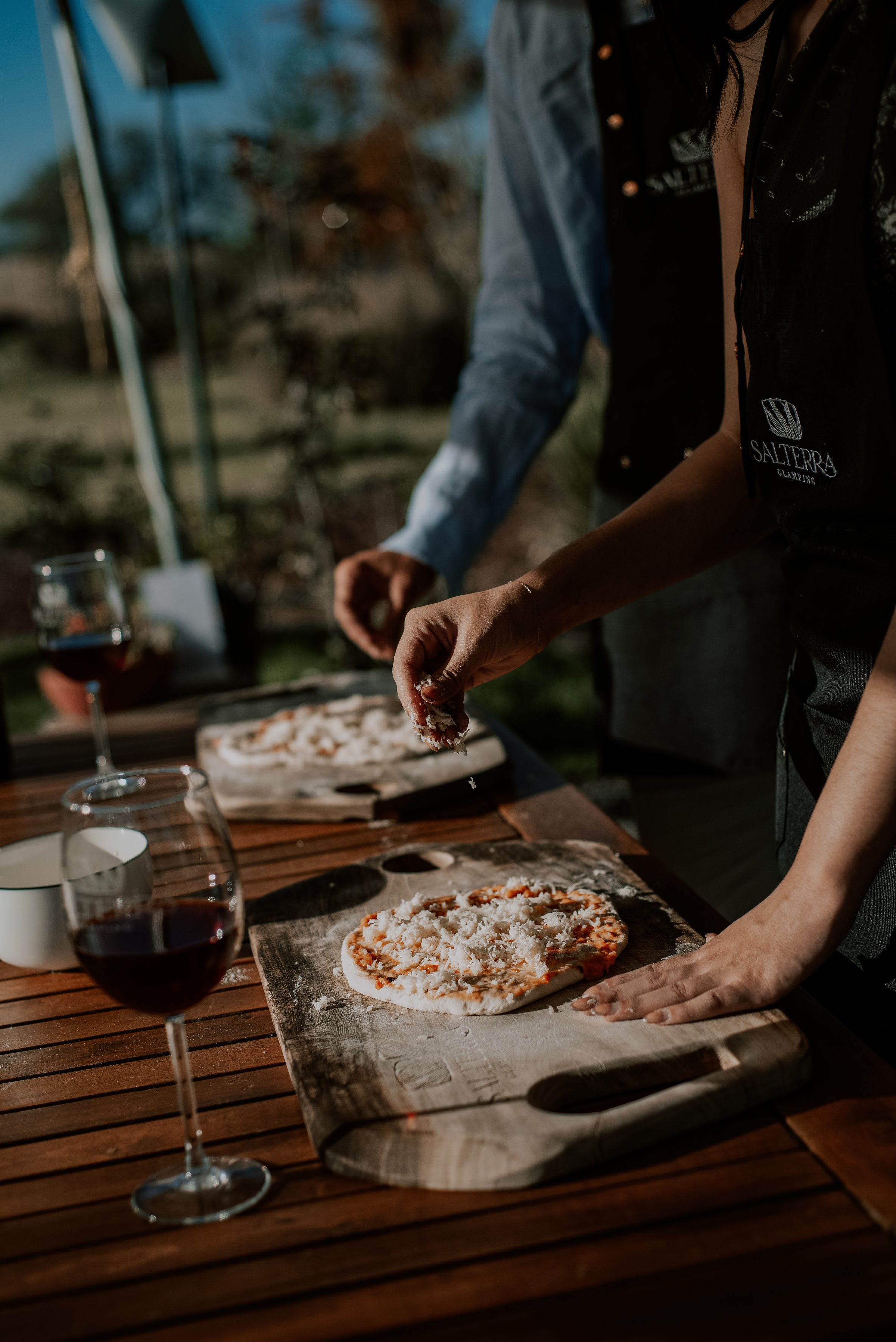 Two people preparing pizzas on wooden boards outdoors, with glasses of red wine nearby on a wooden table.