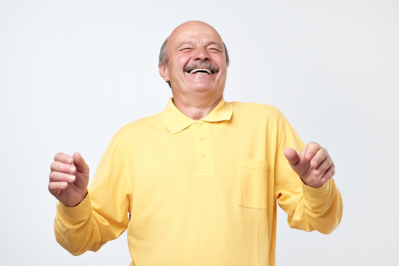 Mature adult man in yellow shirt with moustache laughing looking at the camera over white background. Some jokes are old but gold. Positive emotion concept