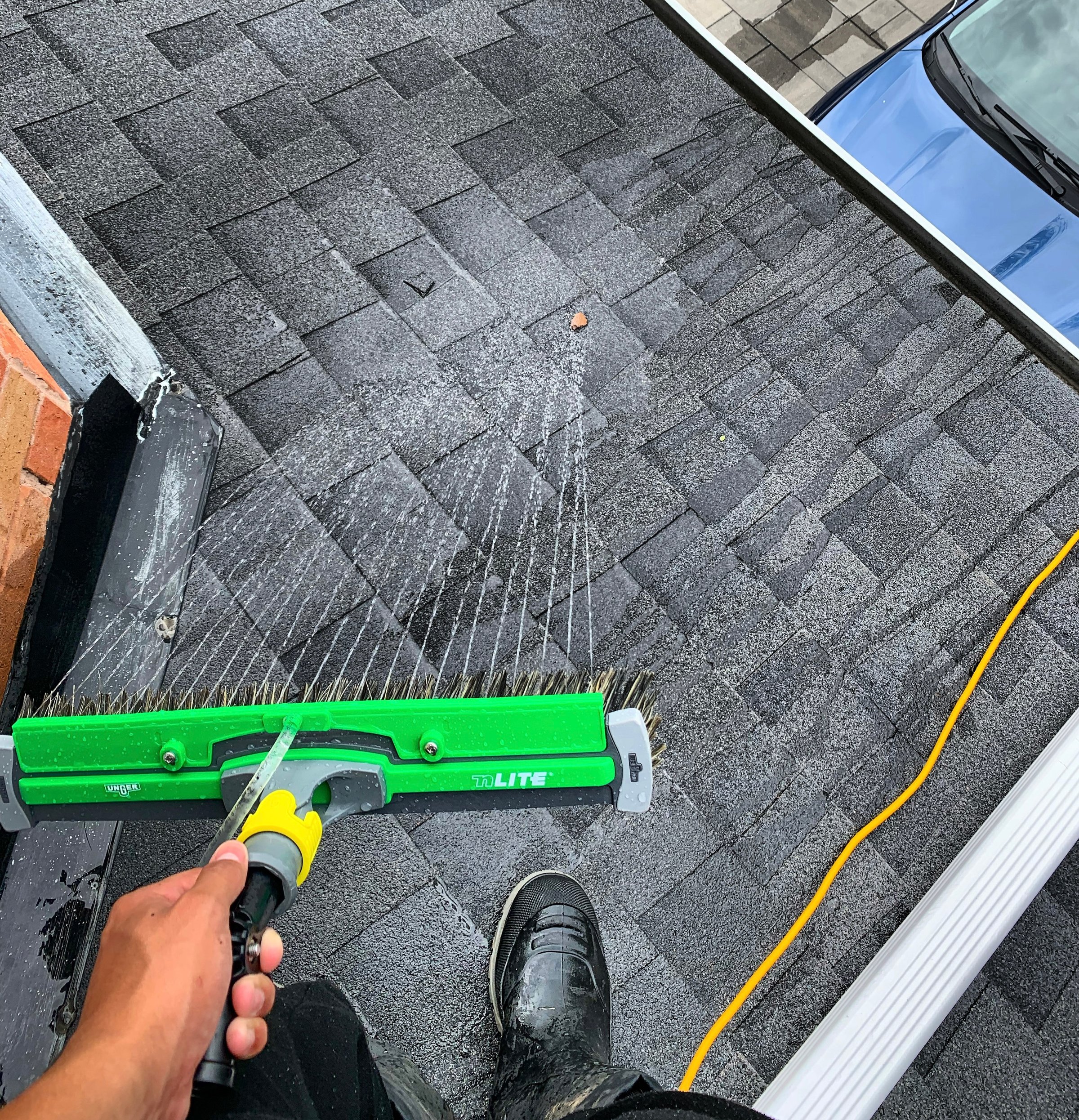 Person cleaning roof shingles with a brush and water spray, standing near gutter. Yellow hose and car visible below.