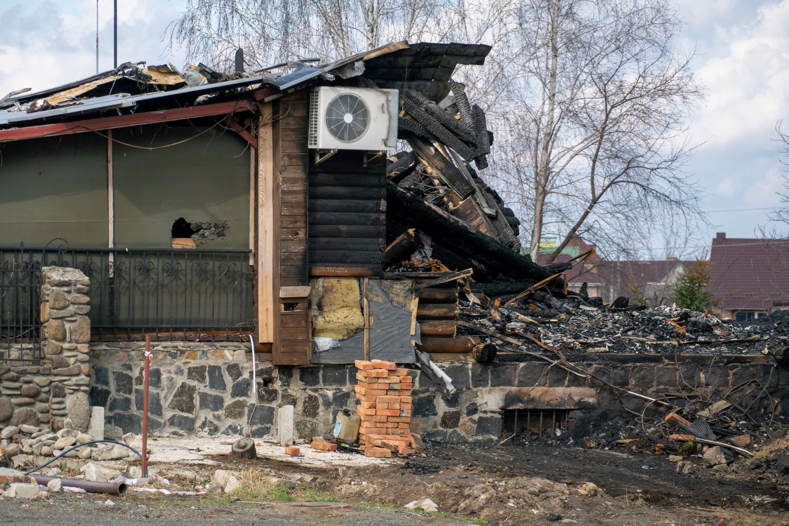 A house stands in ruins following a fierce fire, with charred walls and exposed structures indicating significant damage in a quiet residential area