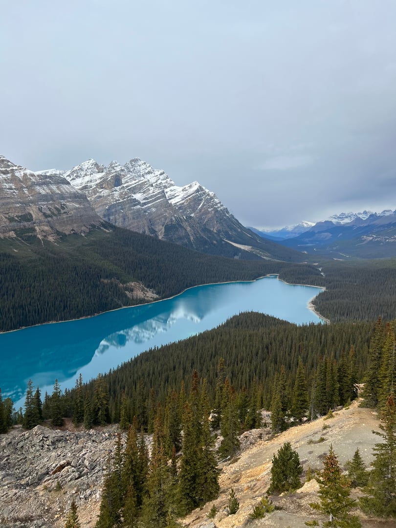 Turquoise lake surrounded by dense evergreen forest and snow-capped mountains under a cloudy sky.