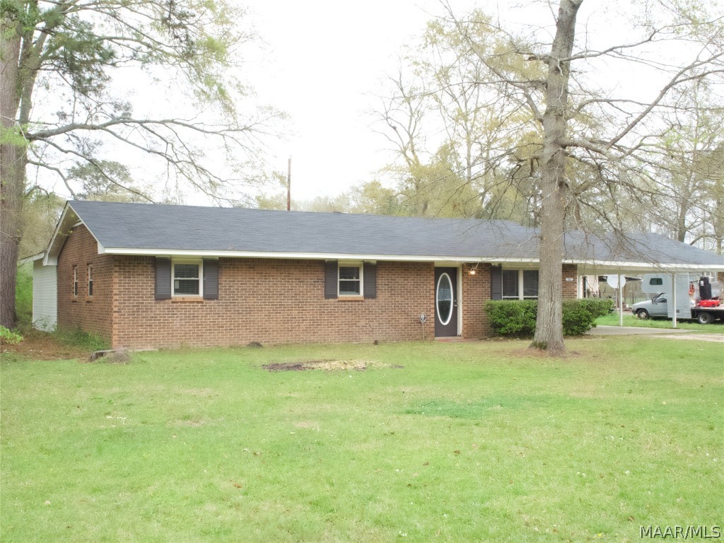 Single-story brick house with a dark roof, front yard, and carport, surrounded by trees.