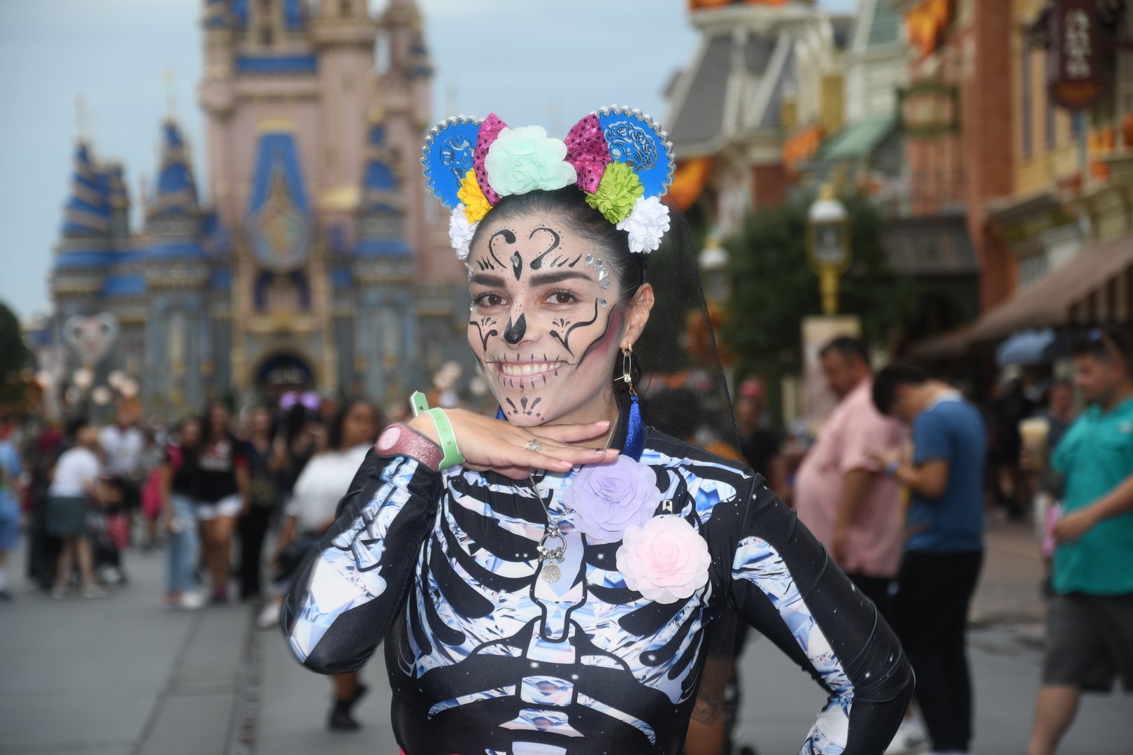 Person in skeleton costume with face paint and flowers poses at a theme park, castle in the background.