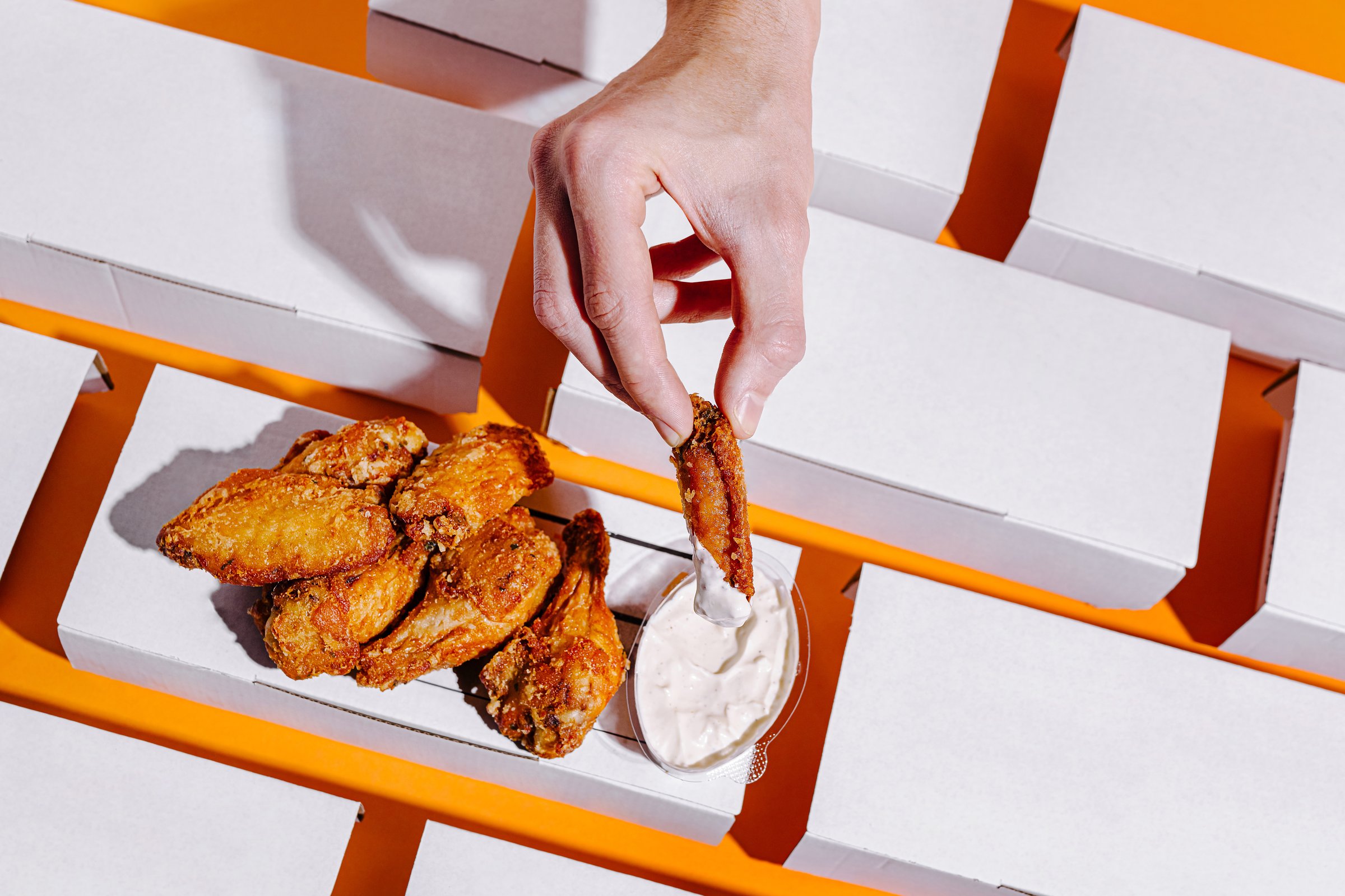A hand reaches into a container, dipping a perfectly fried chicken wing into a rich, creamy sauce. The vibrant orange backdrop contrasts with the white boxes surrounding the delicious meal