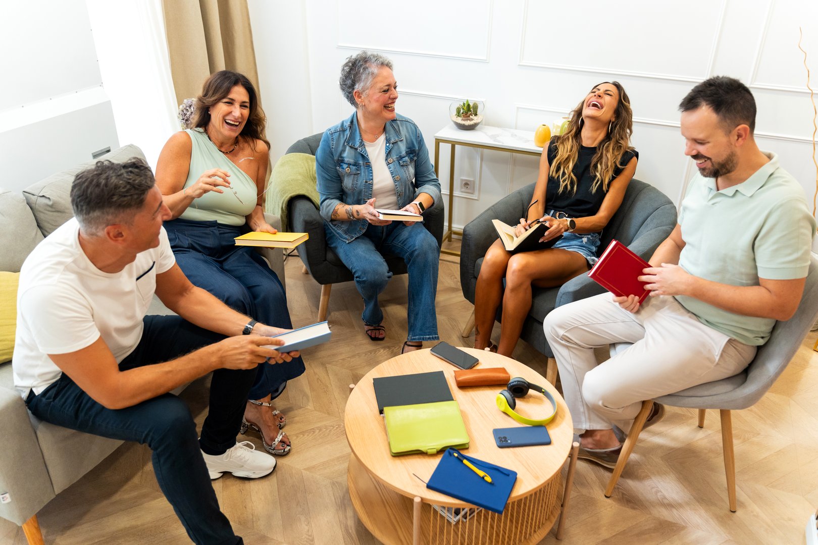 Group of five mixed-age range adults gathering in a living room, holding books and laughing, enjoying a book club meeting