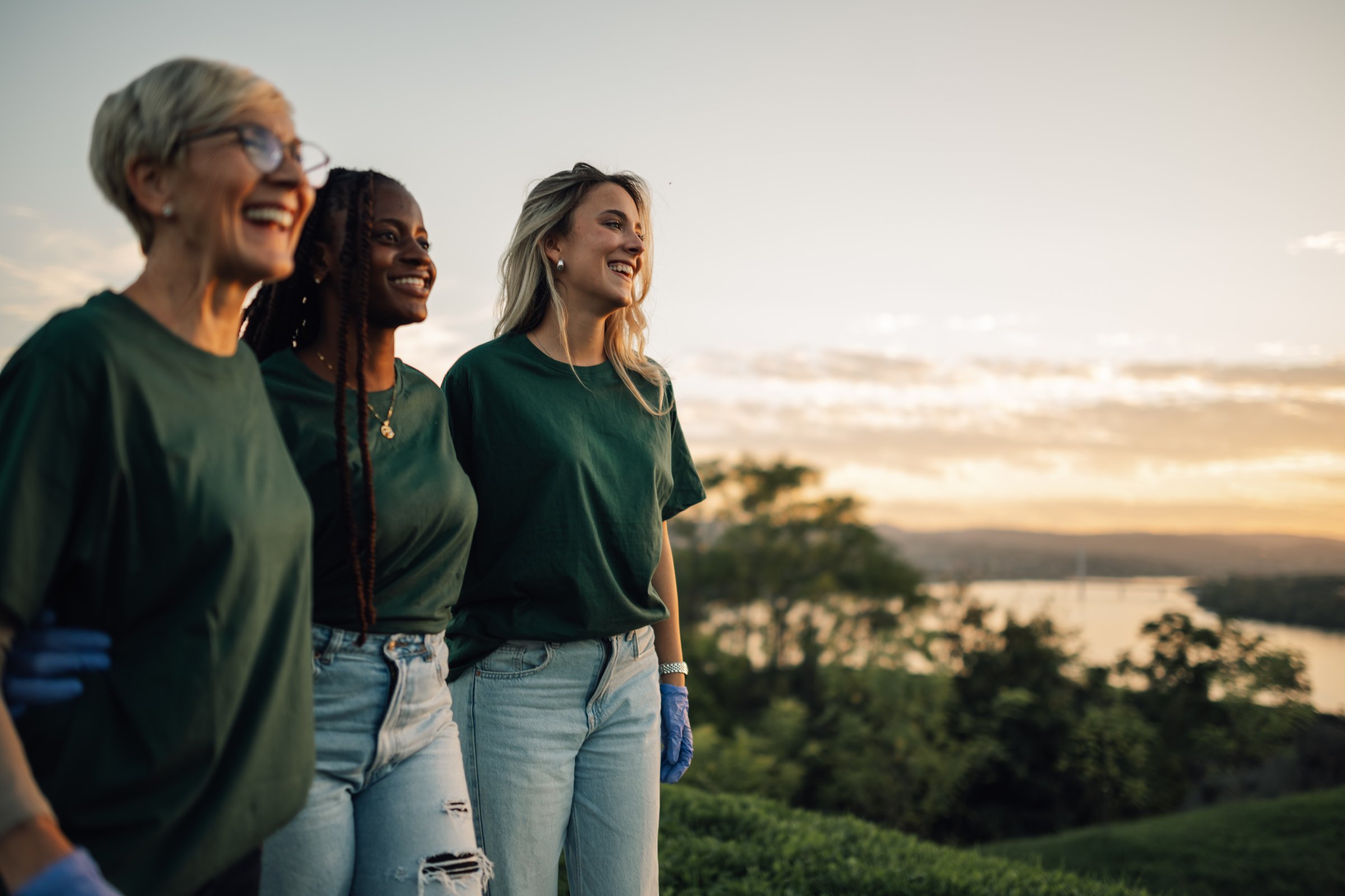 Three volunteers wearing gloves and green t-shirts are walking and admiring the sunset over the river after a rewarding day of environmental cleanup