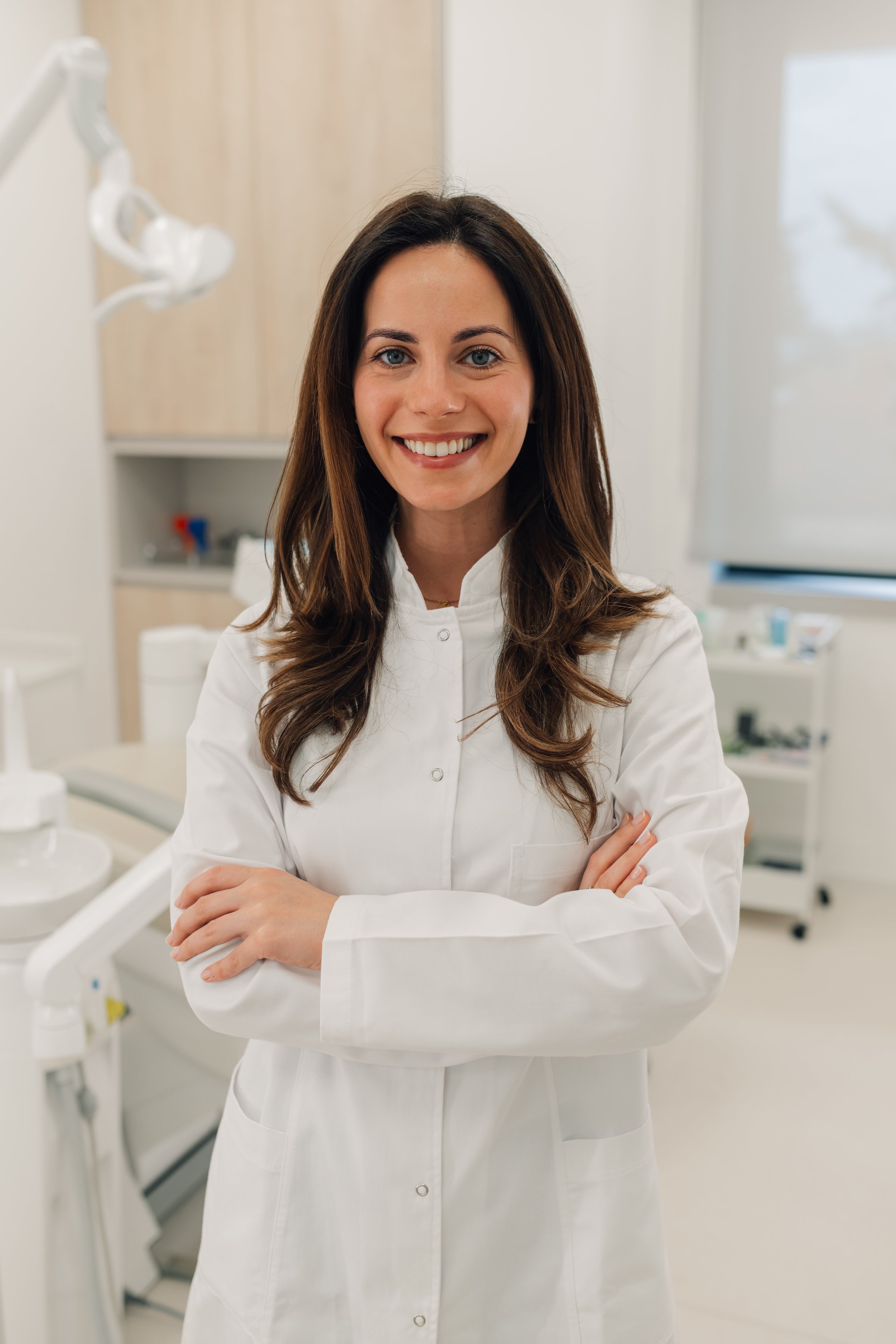 Confident female dentist smiling at the camera with arms crossed, showcasing expertise in a modern, clean dental clinic environment