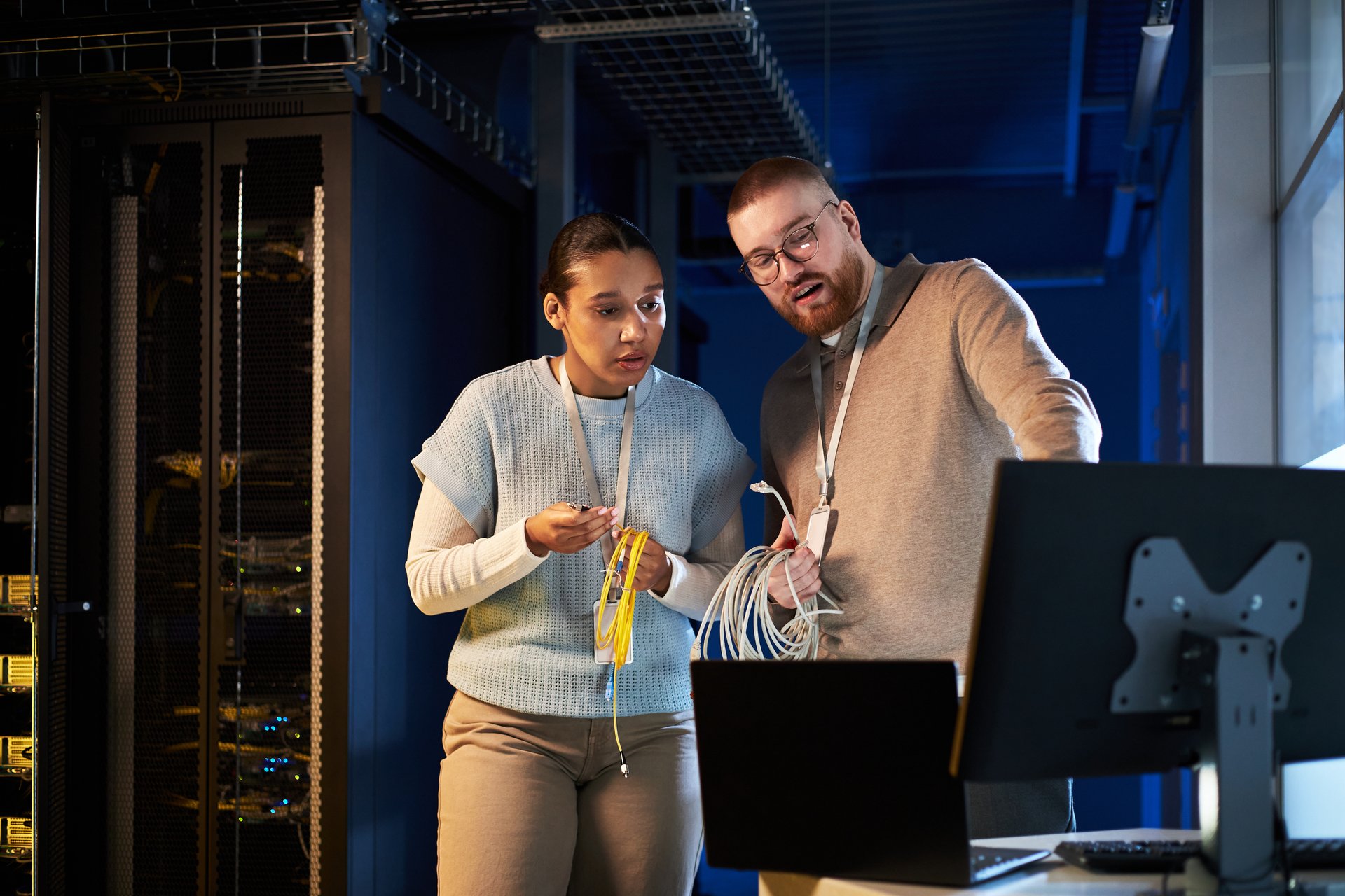 Caucasian female and Caucasian male engineers setting up network infrastructure in modern server room examining cables and hardware with serious expressions