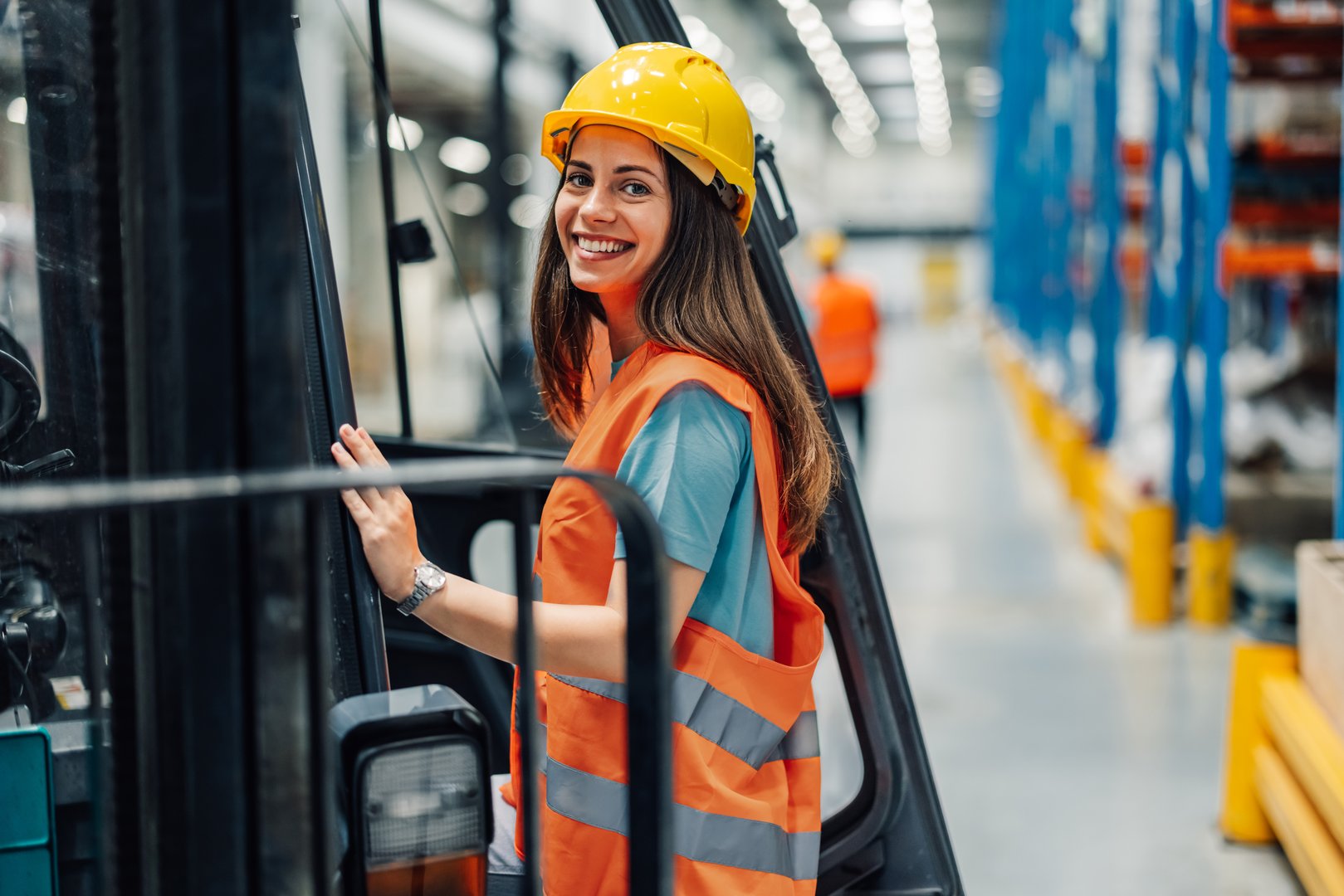 A woman is smiling as she steps into a forklift in a spacious warehouse. She is wearing a yellow hard hat and an orange safety vest, symbolizing a safe work environment.