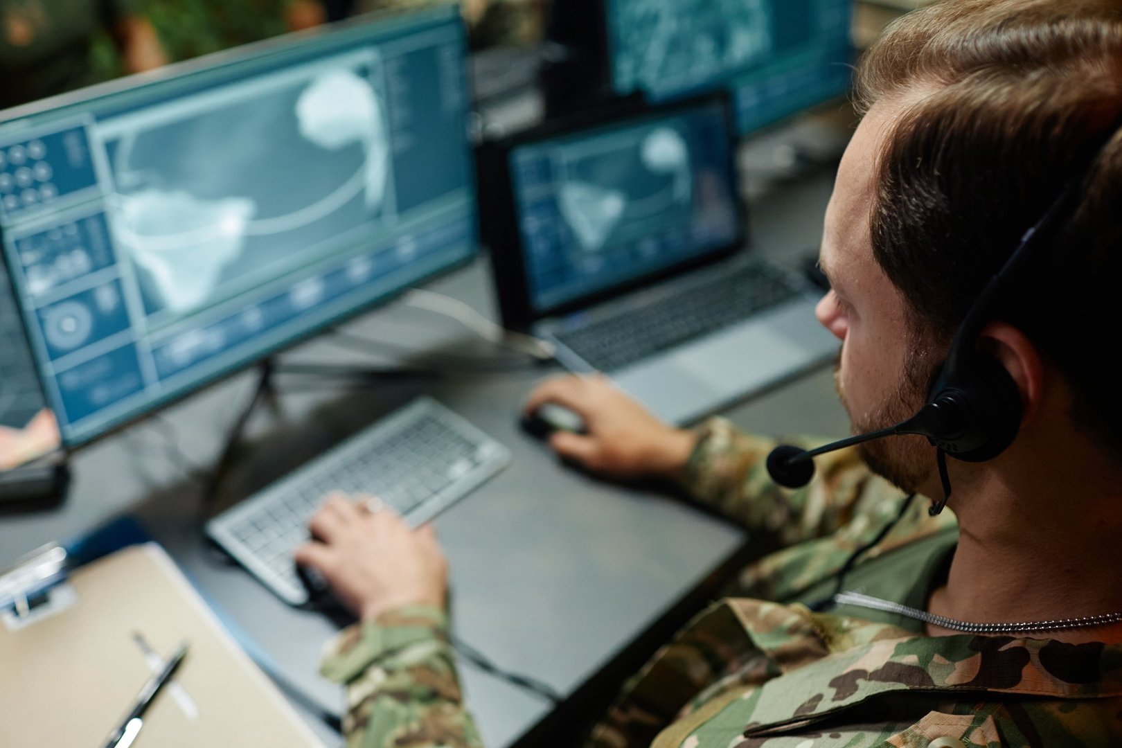 Focus on head of young military officer with headset sitting in front of computer screen with graphic data from surveillance cameras