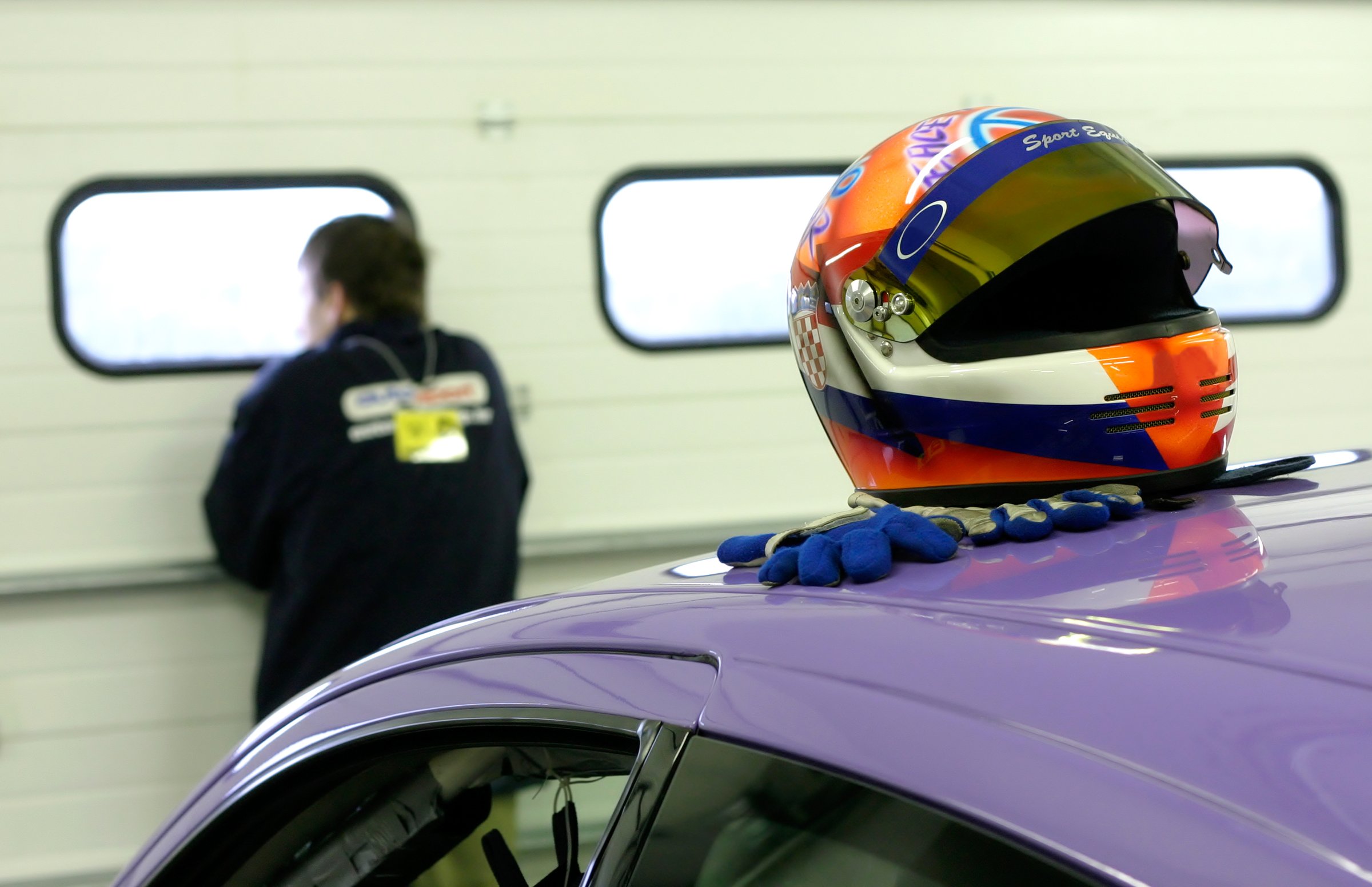 Colorful racing helmet and gloves on a purple cars roof, with a blurred person in the background near garage windows