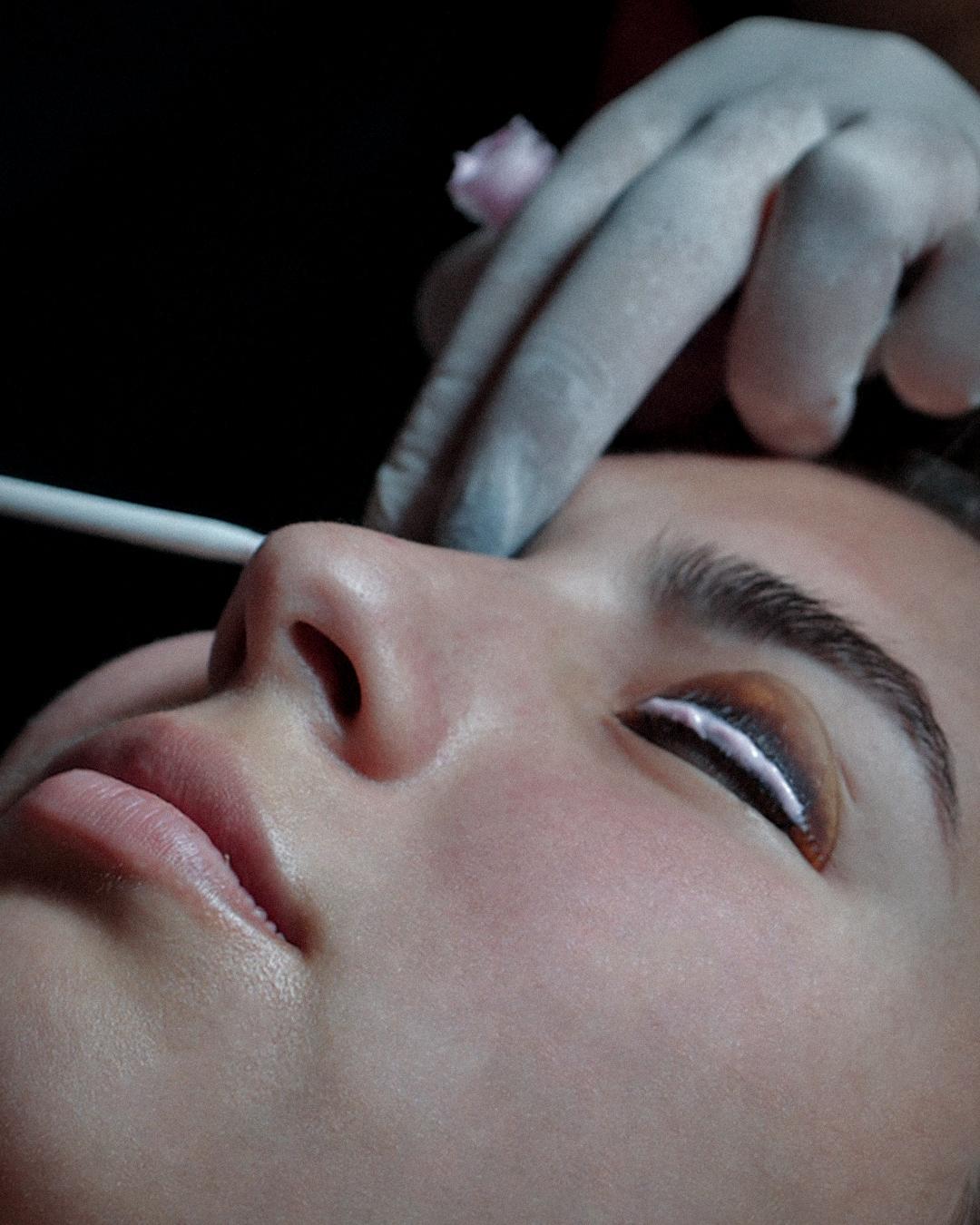 Person receiving an eyelash treatment, with gloved hands applying product near closed eye.