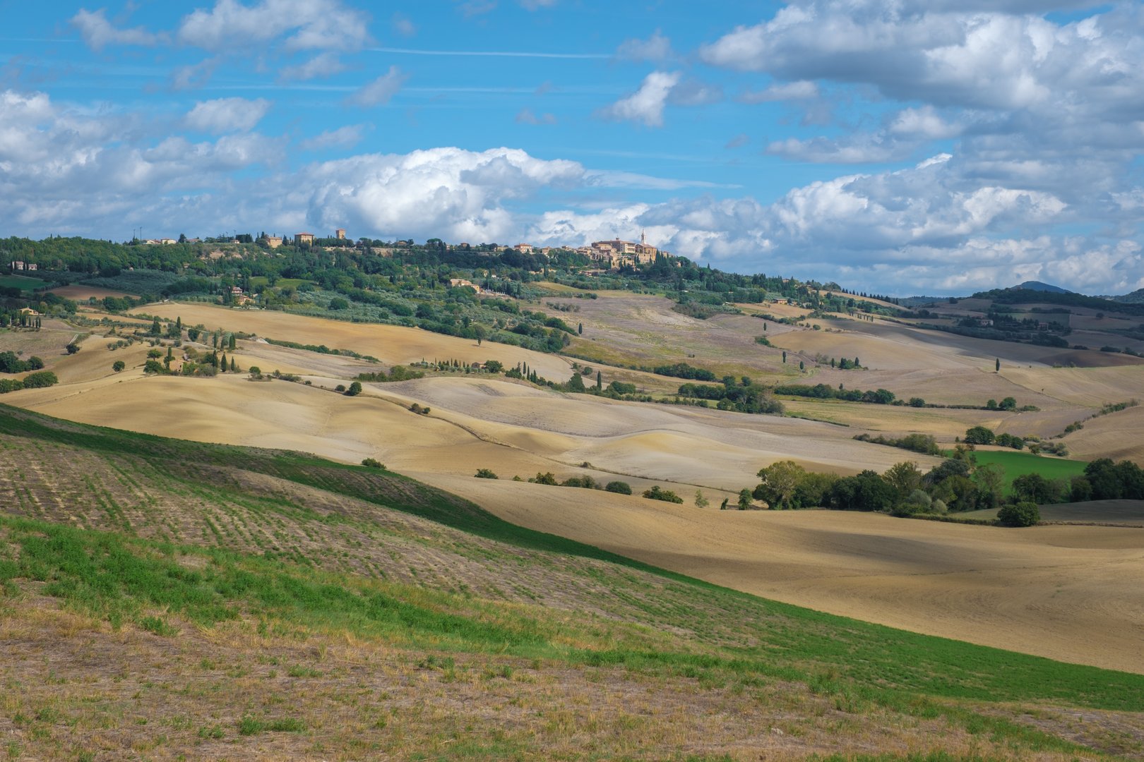 Golden hills stretch as far as the eye can see in Tuscany, Italy, where lush greenery meets soft earth tones beneath a sky dotted with fluffy clouds, creating a serene rural atmosphere.