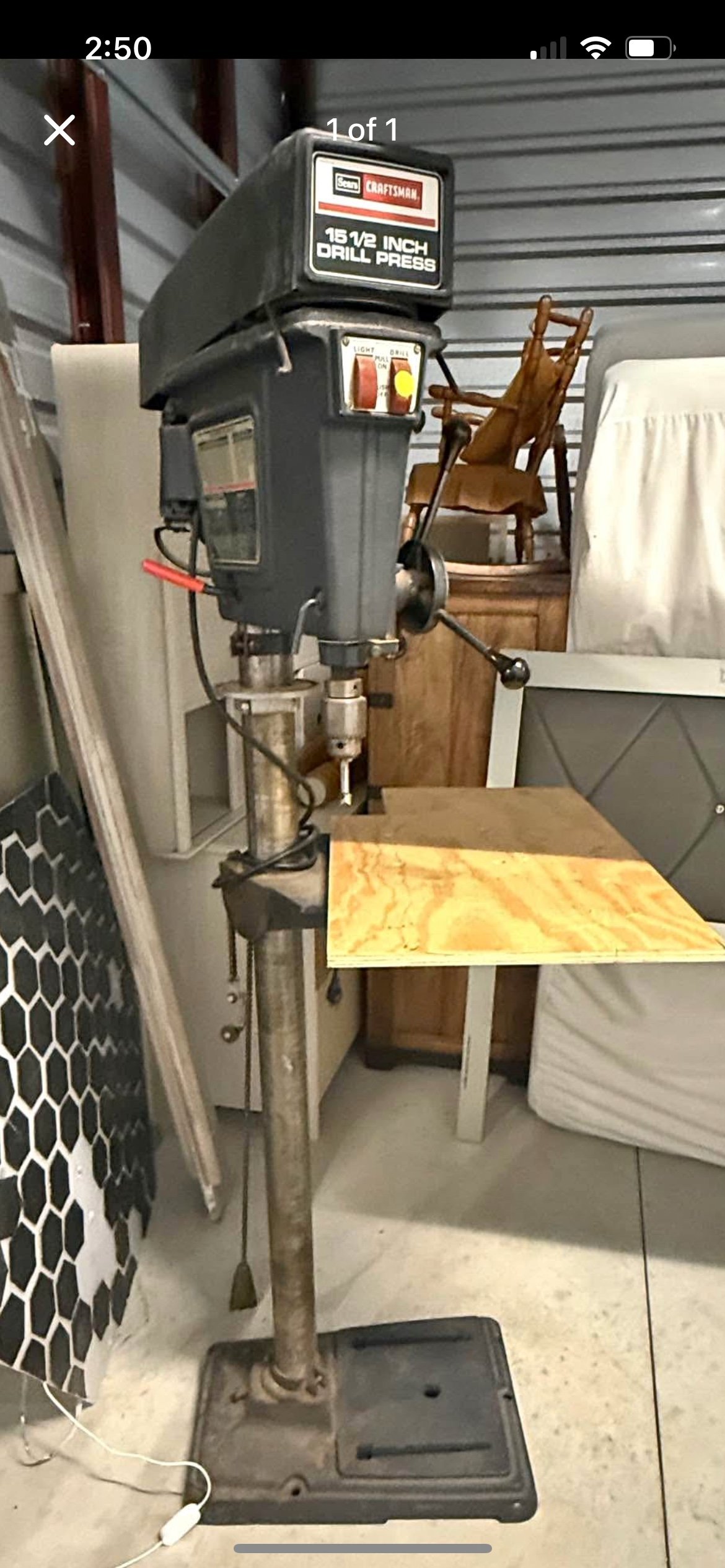 A Craftsman 15 1/2 inch drill press with a wooden board clamped to it, in a garage with furniture and various items nearby.