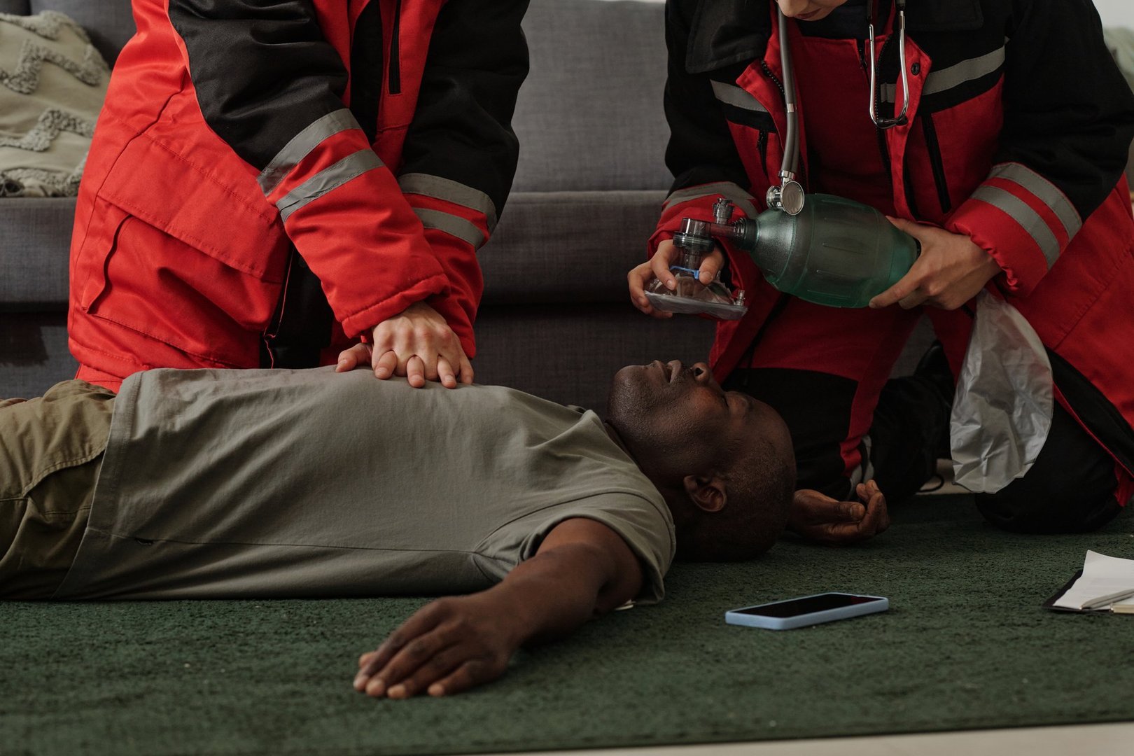 Paramedics attending to unconscious man on floor with medical equipment in hands. Scene shows emergency response team working for patient on green carpet near couch with cellphone nearby