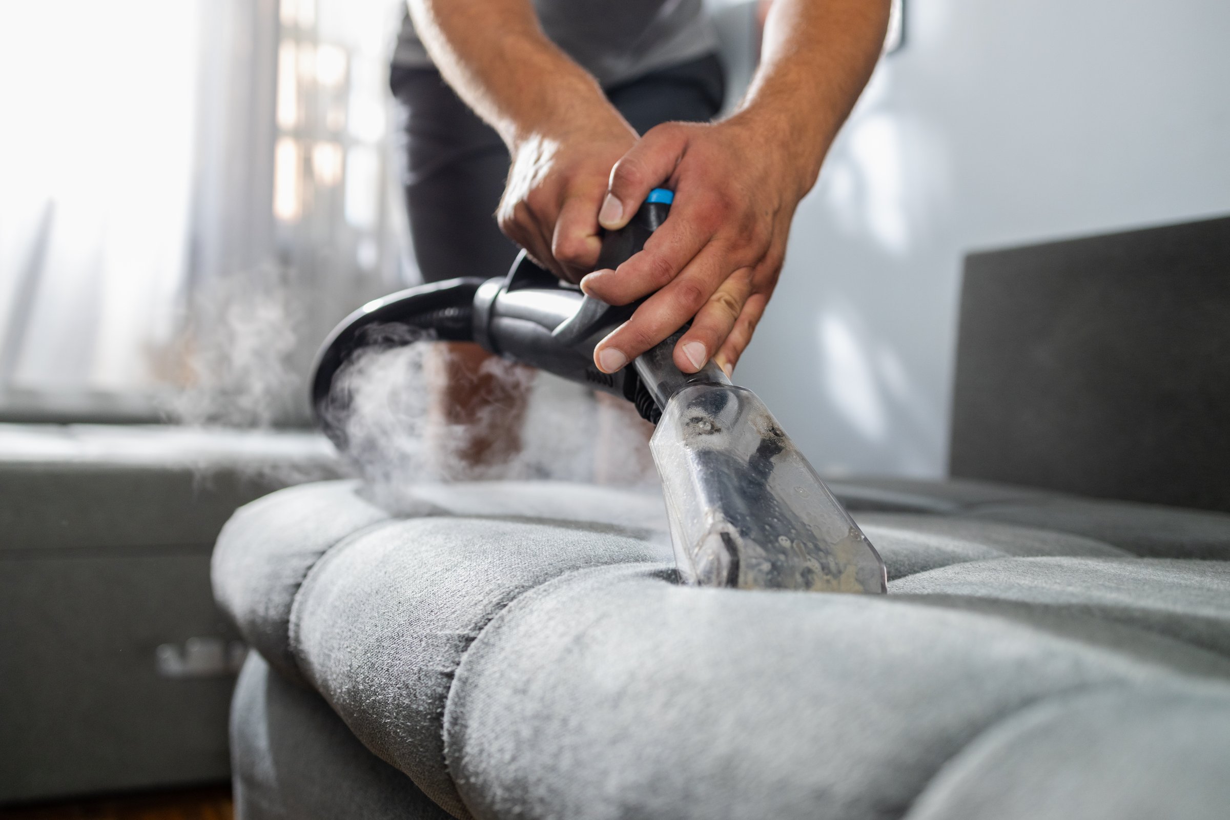 Man's hands operating a steam cleaner, deep cleaning and sanitizing sofa upholstery