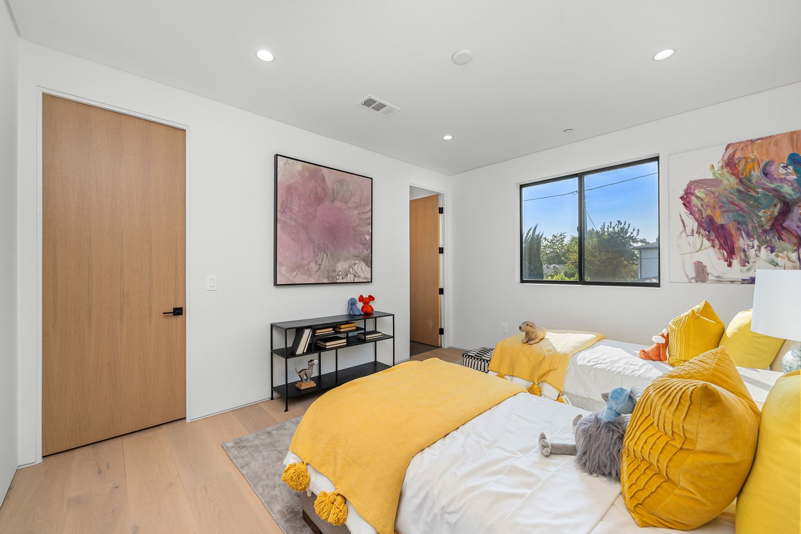 Modern bedroom with twin beds, vibrant yellow accents, and abstract art, featuring natural light through a large window.