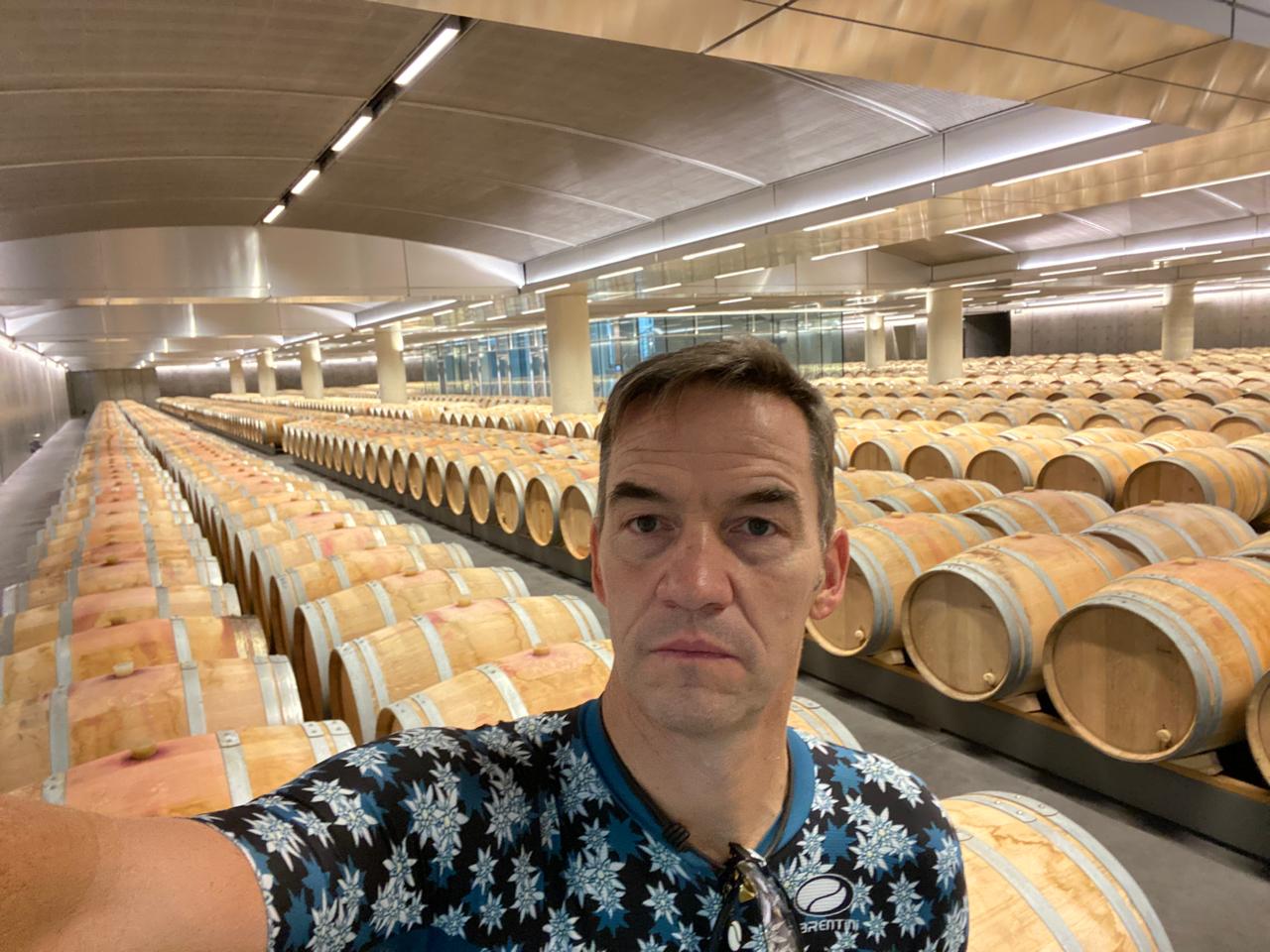 Man taking a selfie in a large wine cellar with rows of wooden barrels.