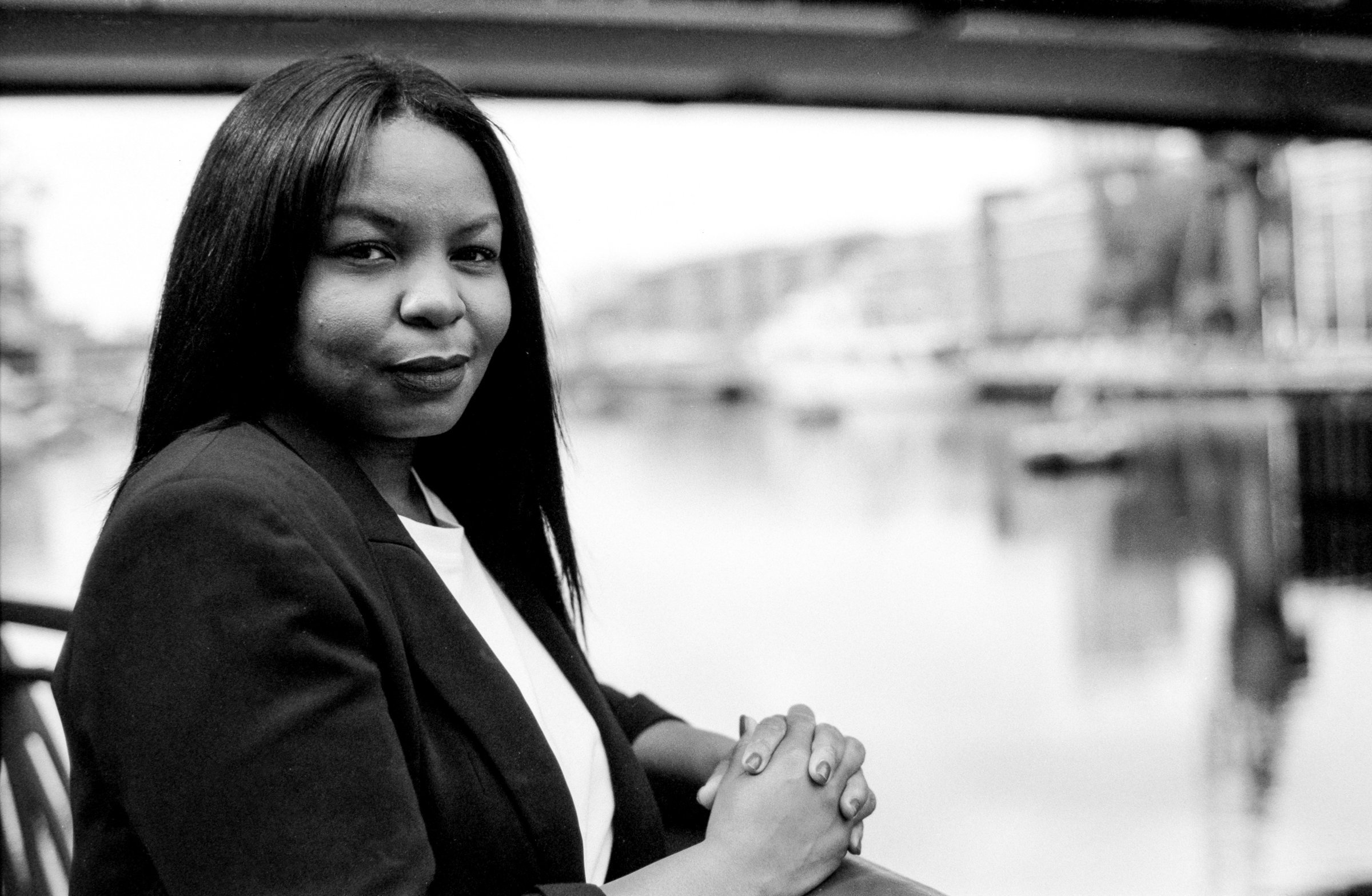 Grainy monochrome analog image depicting a confident businesswoman posing near a canal in an urban setting, with her hands clasped and a slight smile