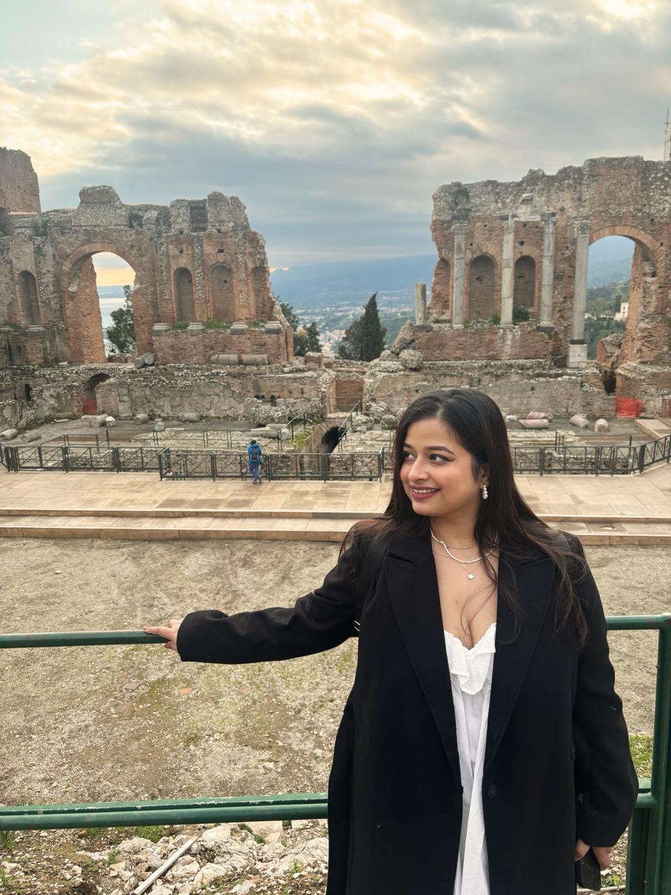 Woman in a black coat stands in front of ancient ruins under a cloudy sky, smiling, with an expansive view in the background.
