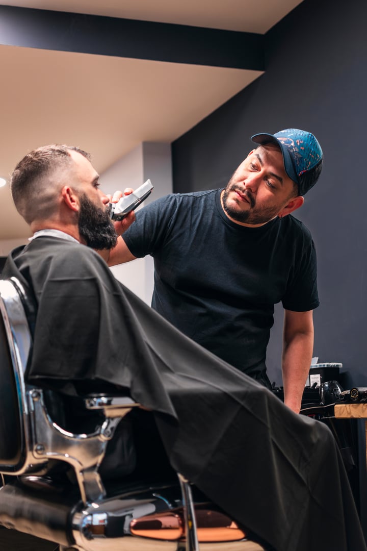 A man is having his beard trimmed with an electric razor. The man is sitting in an old barber's chair. The barber is wearing a black shirt and a baseball cap. The image is casual
