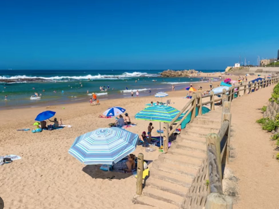 A sandy beach with people sunbathing under umbrellas and swimming in the sea, featuring wooden stairs leading to the shore.