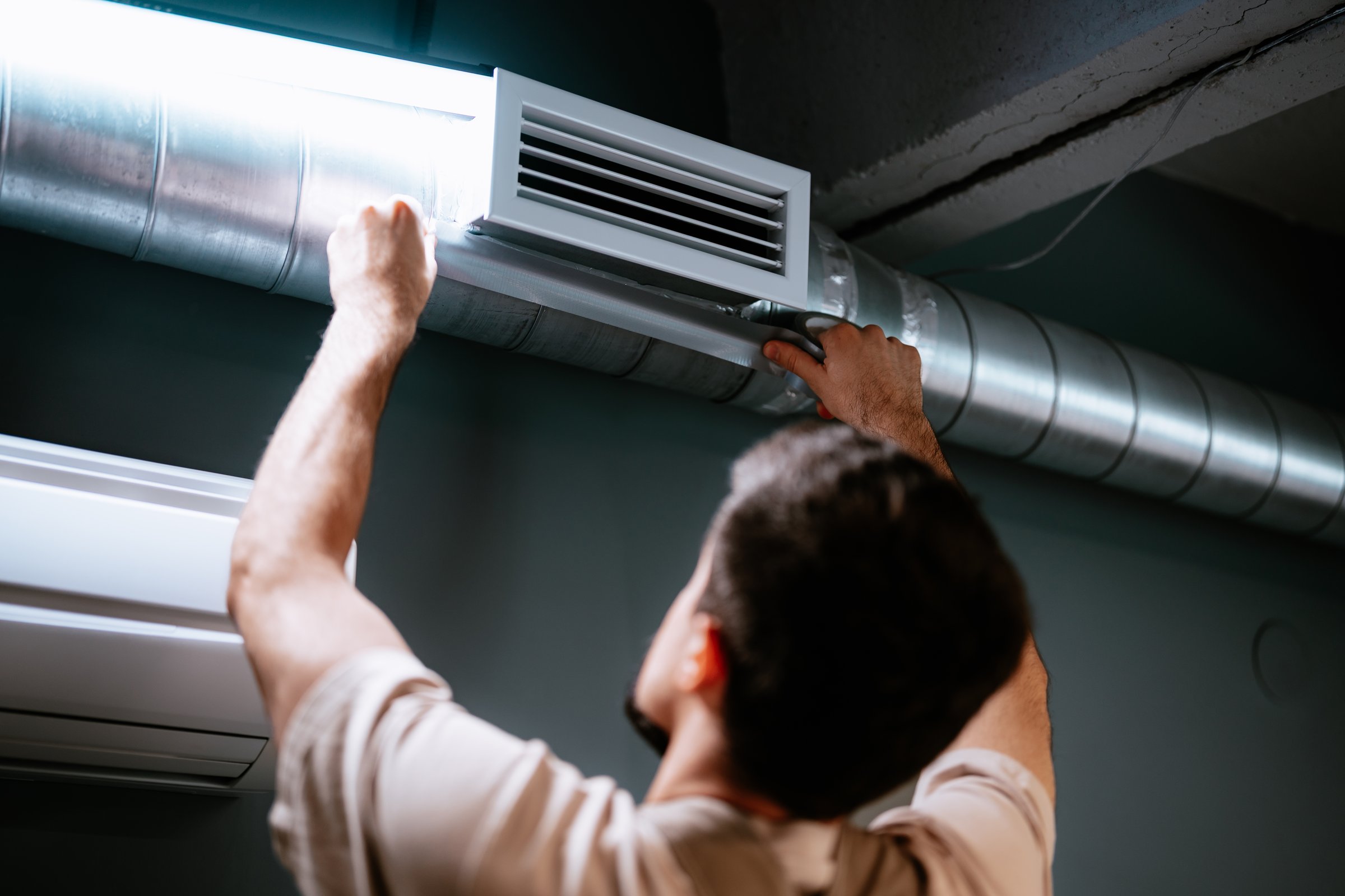 A technician is adjusting an air duct system in a contemporary space. The focus is on improving air circulation. Tools are visible, and the setting is well-lit with metallic elements.