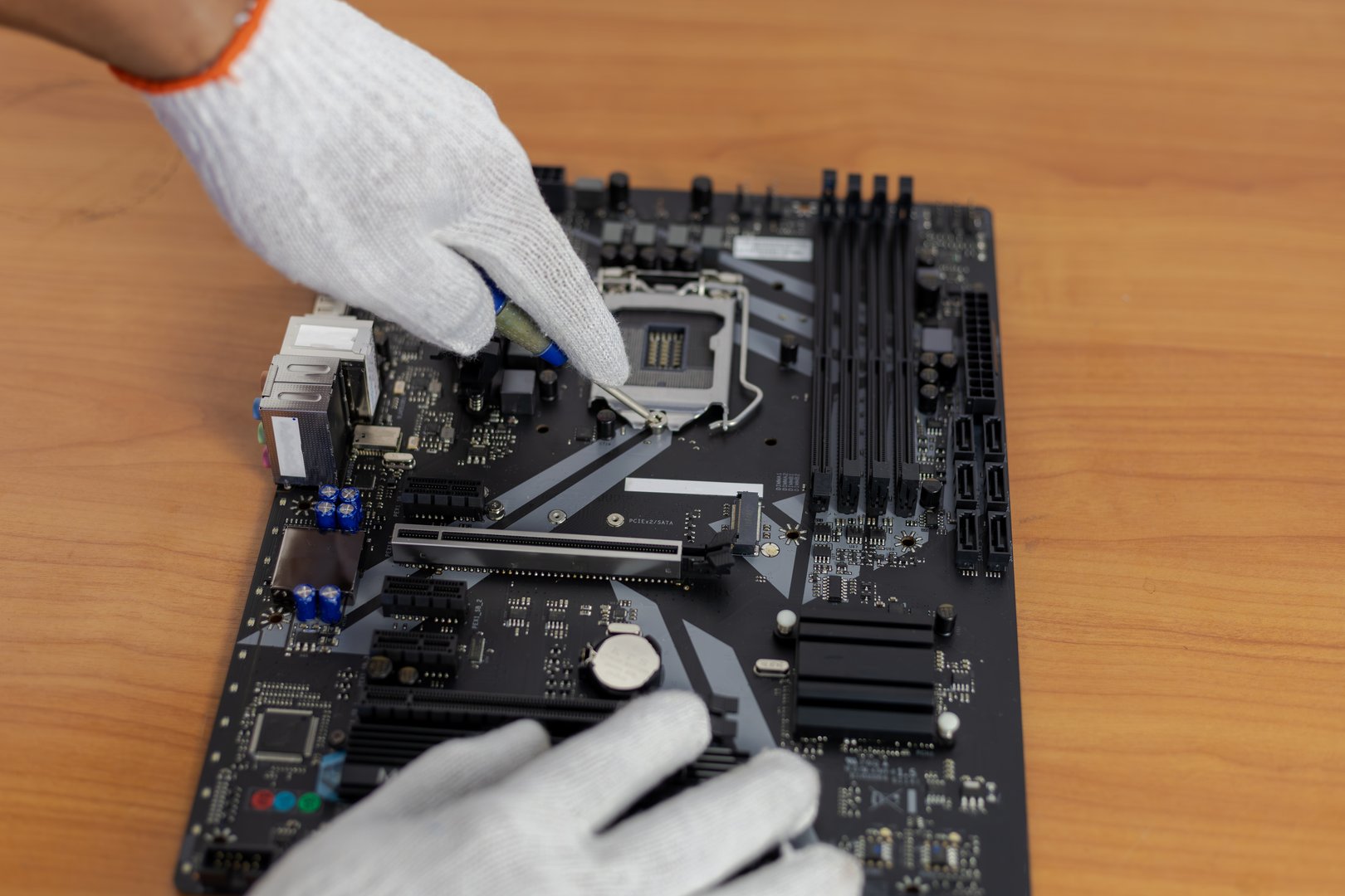 A close-up shot of gloved hands using a tool to work on the CPU (Central Processing Unit) socket area of a black computer motherboard.