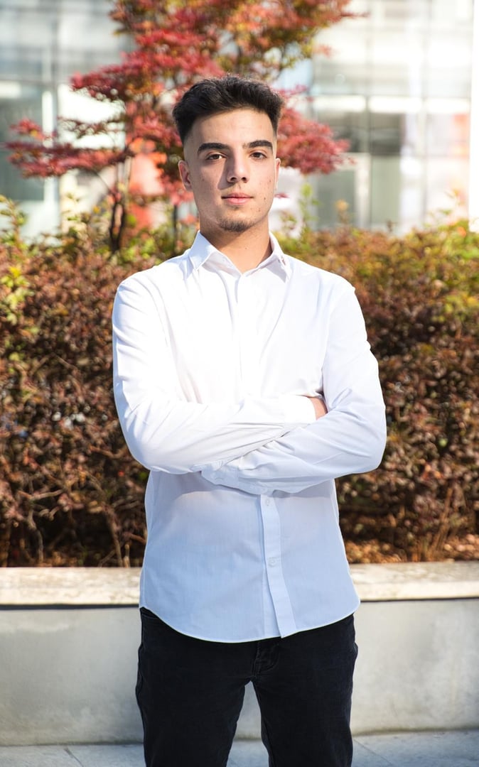 A young man in a white shirt standing in front of a stone wall, looking at the camera.