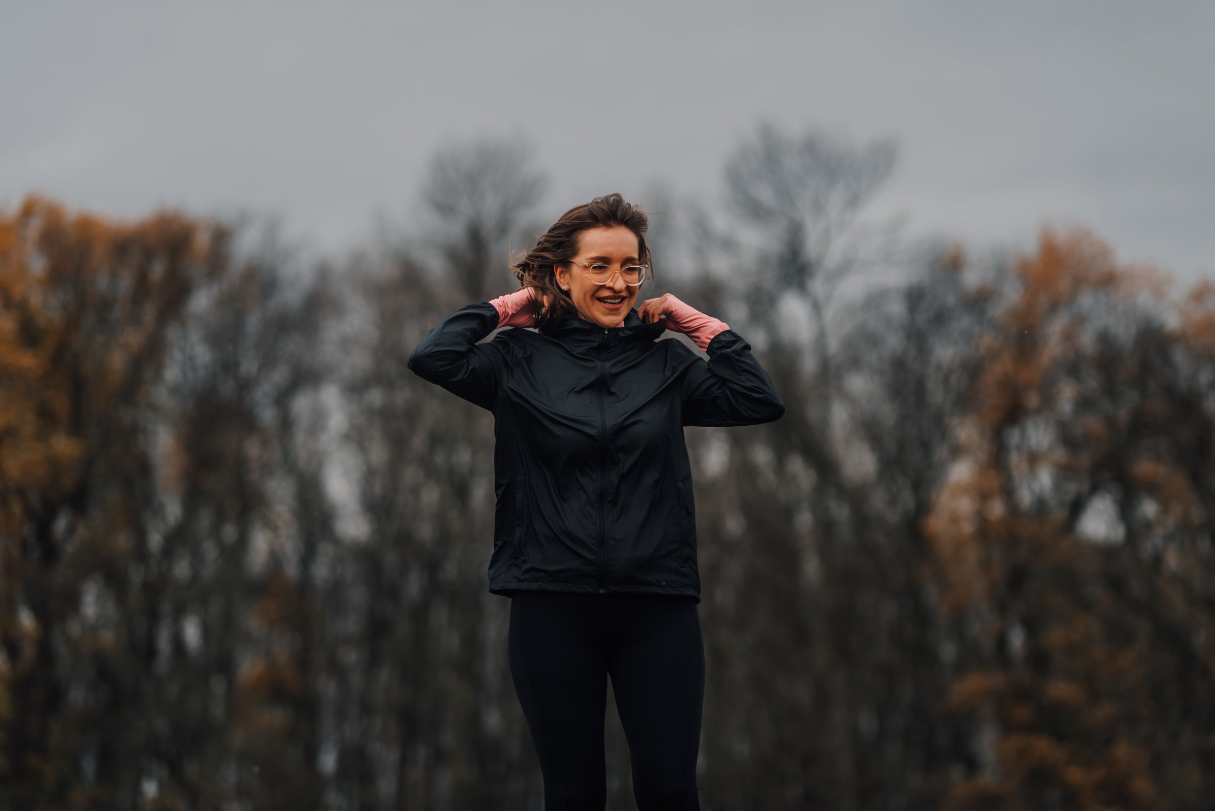 Smiling sportswoman putting on the hood of her black jacket in a park during a cloudy autumn day