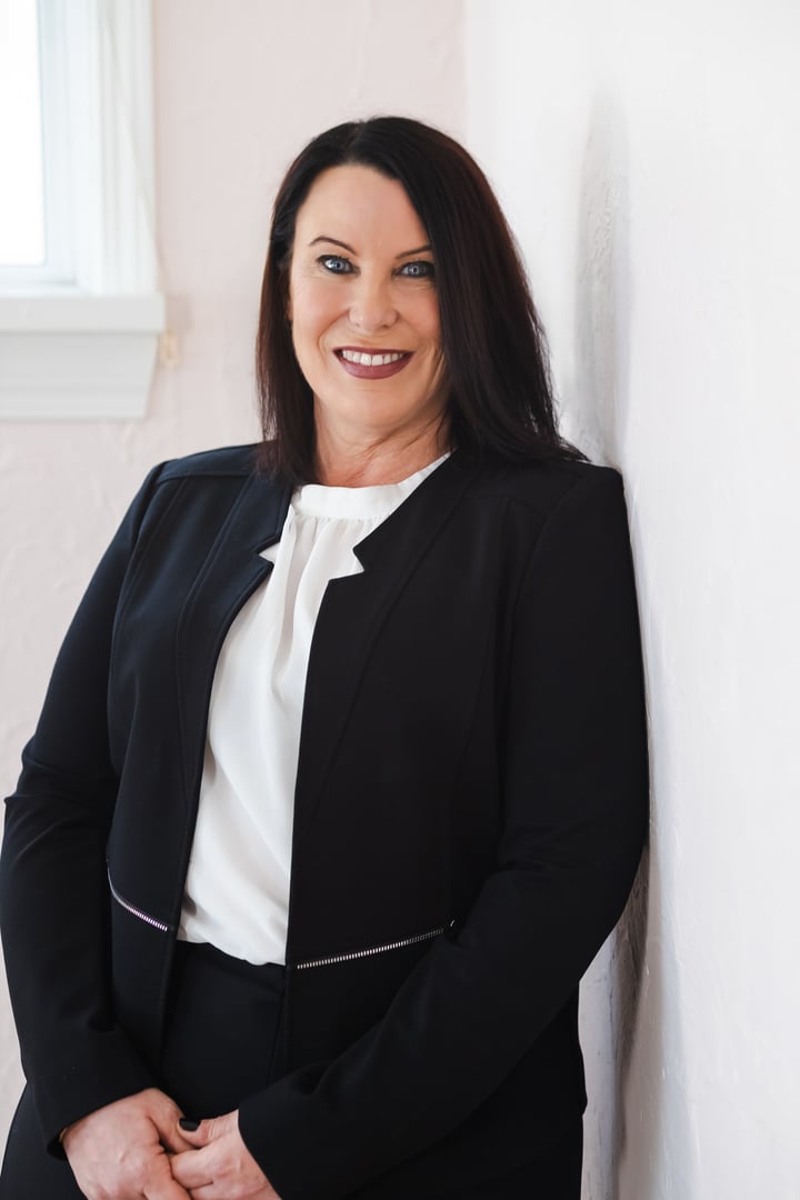 Woman in a black blazer standing against a white wall, smiling.