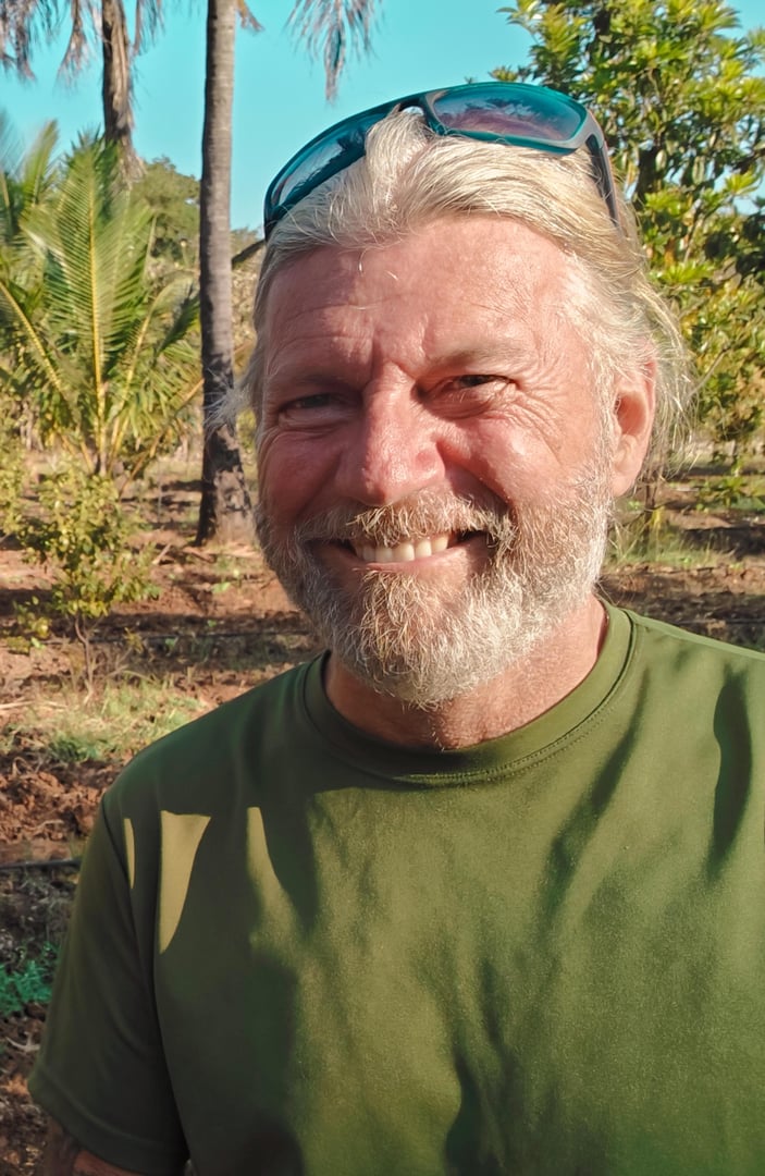 Man with gray beard and sunglasses on head smiling outdoors in a tropical setting with palms in the background.
