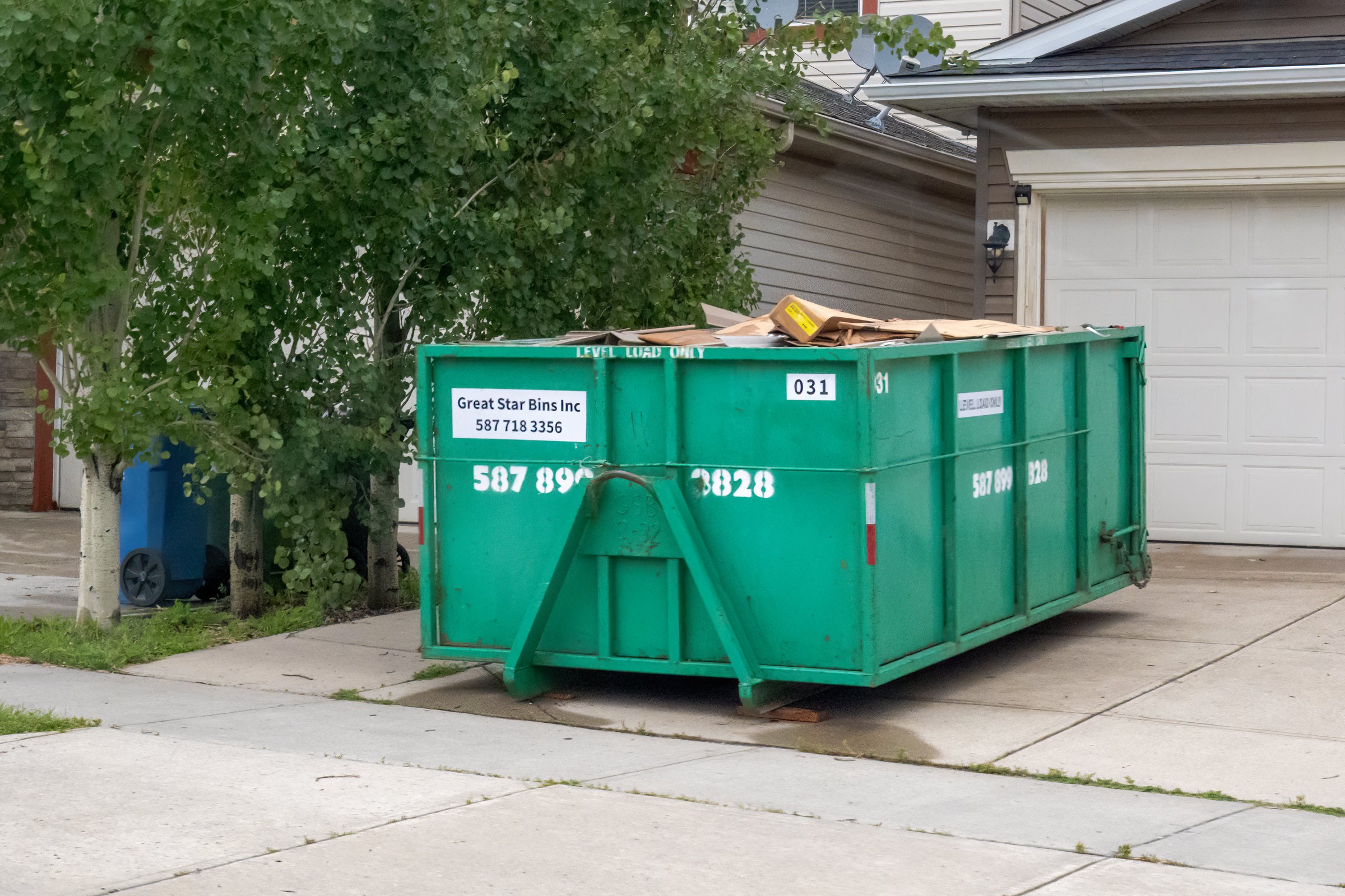 Calgary, Alberta, Canada. Oct 28, 2025. A large green Great Star Bins roll-off dumpster, filled with debris, parked in a residential driveway for waste removal.