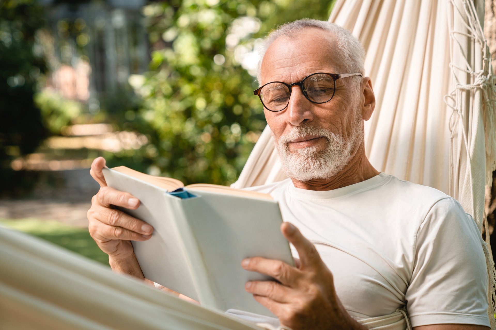 Concentrated dreamy old senior elderly caucasian grandfather man relaxing resting in hammock while reading book outdoors in park garden forest