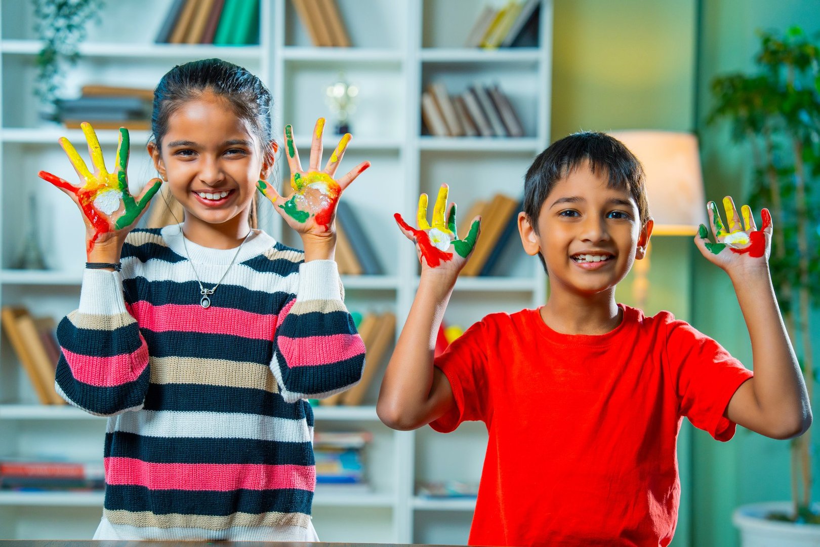 Cute Little Playful Indian Asian Siblings Showing Both Palms Covered with Bright Colors, Smiling Joyfully, and Expressing Creativity in a Fun Indoor Painting Activity at Their Modern Home