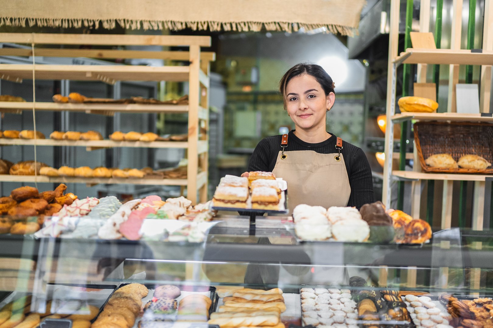 Young saleswoman arranging delicious pastries in a bakery shop display case, smiling at the camera