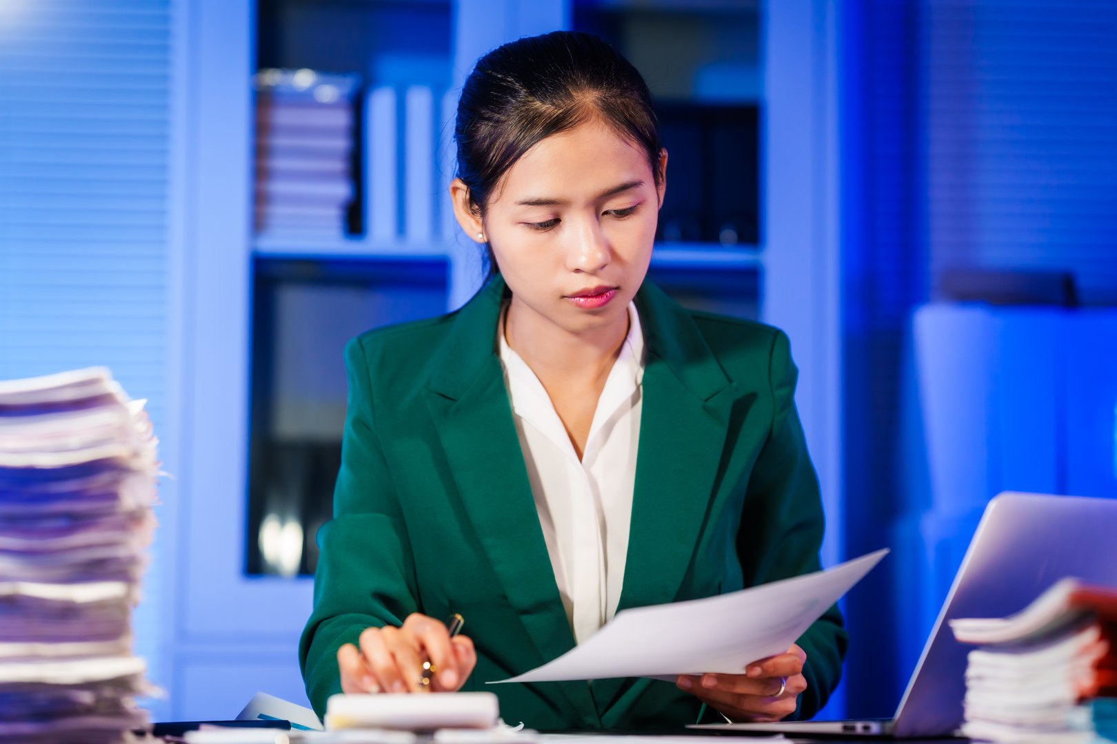 An Asian businesswoman works overtime,sitting at her desk at night, searching through numerous documents on her PC or laptop.Facing office problems,she also chats online while handling lots of work