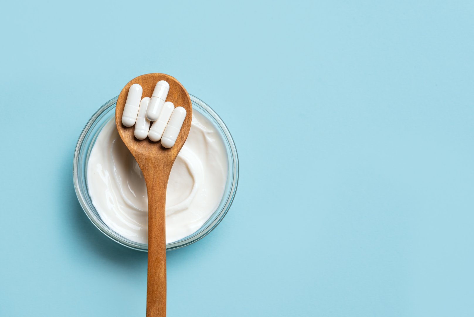 Probiotic capsules on wooden spoon and bowl of kefir or yogurt on blue background, top view, copy space. Fermented organic foods, nutritional supplement, healthy diet for digestion concept.