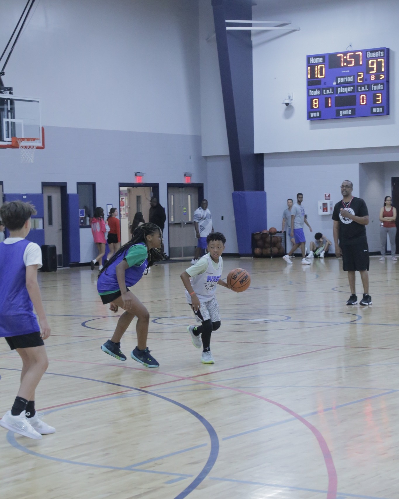 Youth basketball game in a gym. A young player dribbles the ball while others guard. Scoreboard and coach visible.