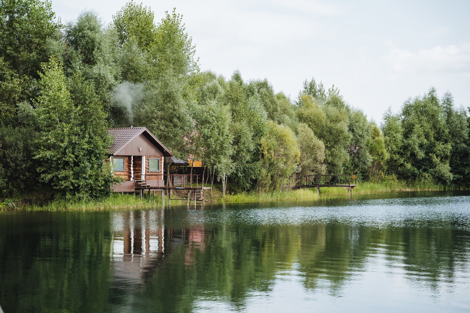 A quaint, small wooden house is peacefully sitting on the edge of a beautiful lake, which is surrounded by tall trees and lush greenery, creating a serene and picturesque natural landscape