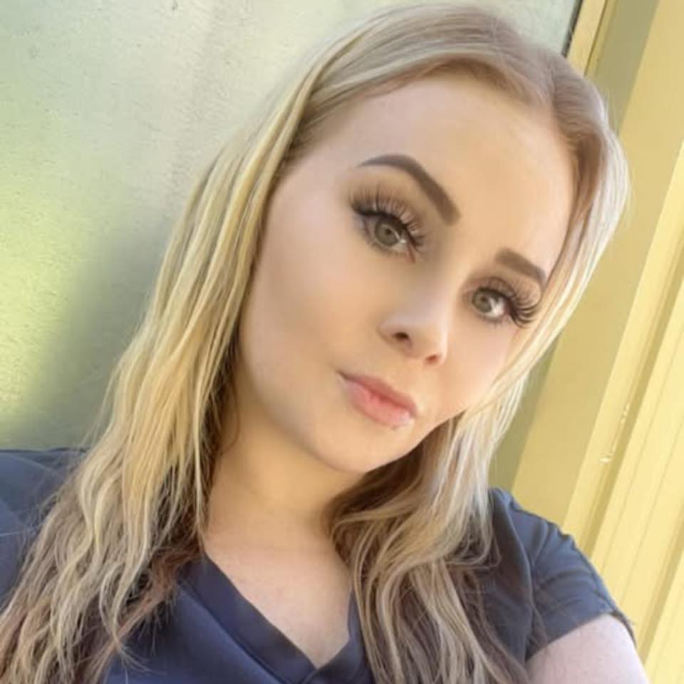 A woman with long blonde hair and wearing a dark top poses for a selfie against a light-colored background.