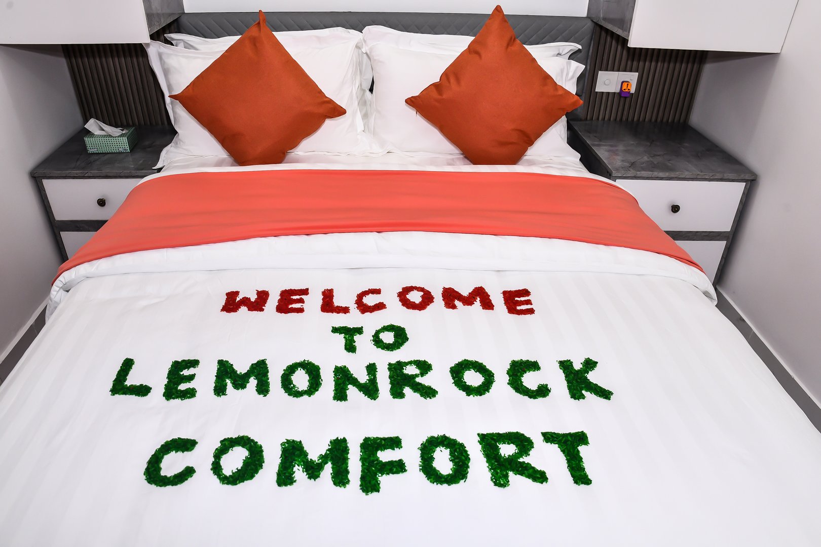 A neatly made bed with a message "Welcome to Lemonrock Comfort" arranged on the blanket using green and red materials.