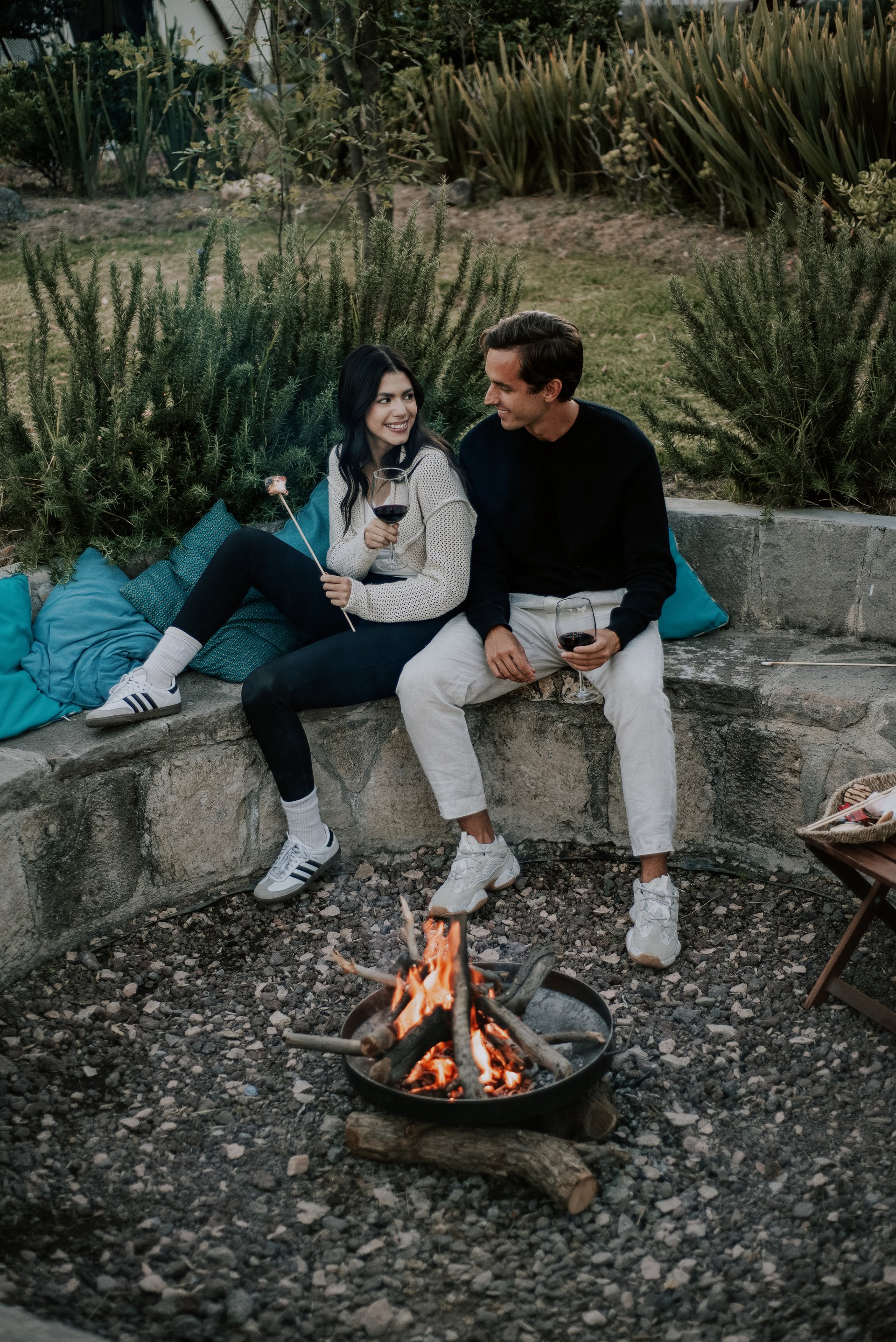 A couple sitting by a fire pit, holding drinks, and smiling at each other. They are outdoors on stone seating with cushions.