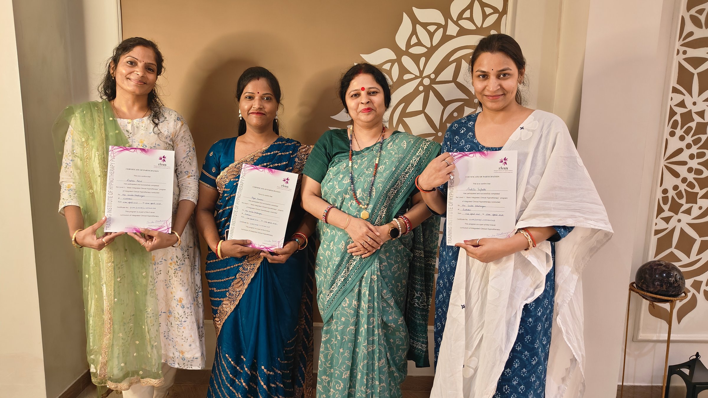 Four women in traditional attire, two holding certificates, pose for a photo against a decorated backdrop.