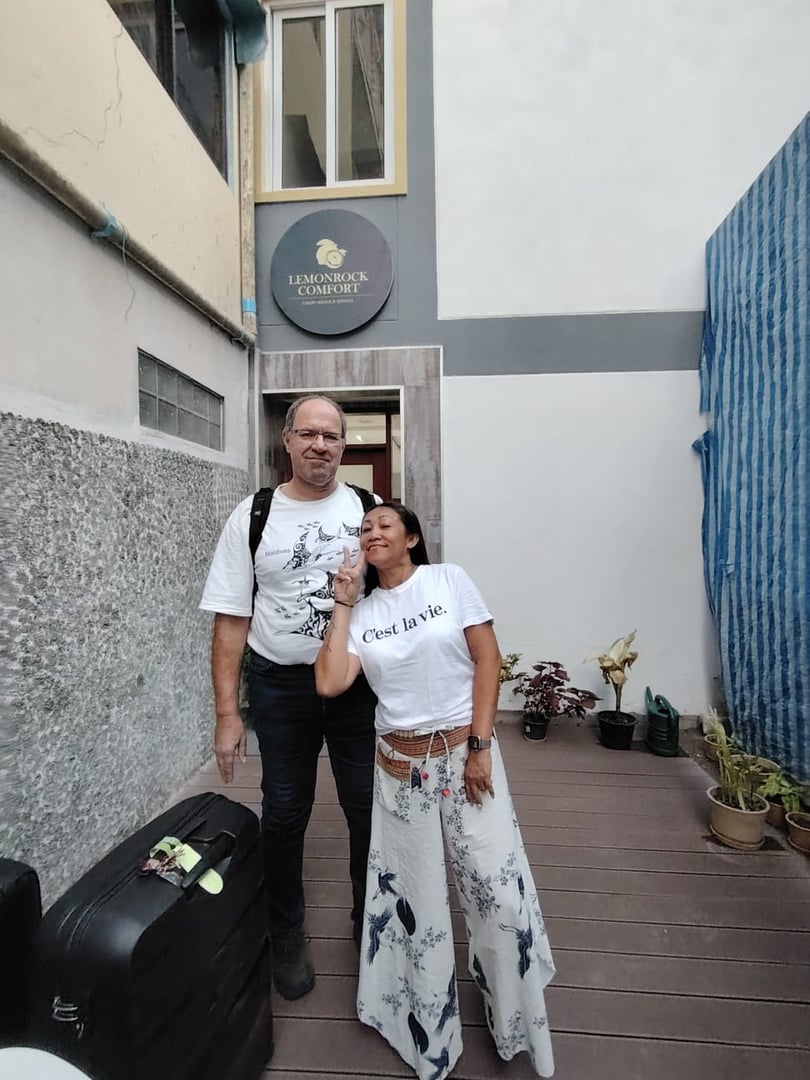 A couple standing with luggage outside "Lemonrock" hotel, both wearing casual summer outfits, smiling at the camera.