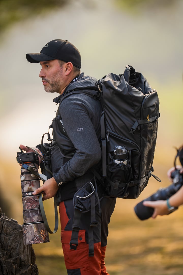 Man with a large backpack and camera gear, wearing a cap and outdoor clothing, stands in nature, ready to photograph.