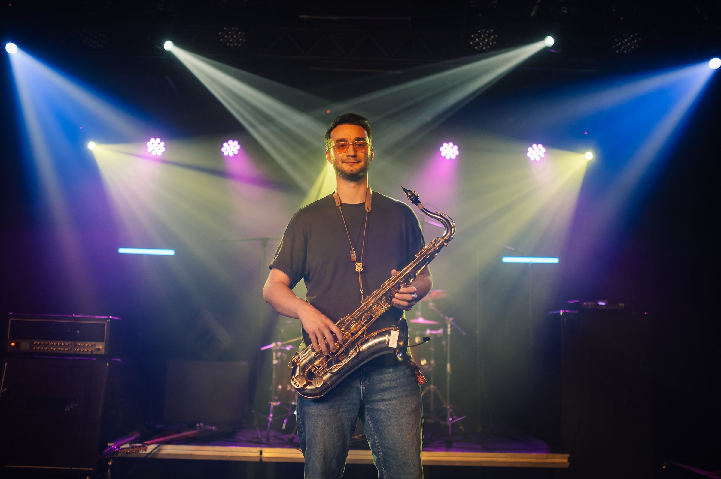 Young male musician holding a saxophone, standing on a stage illuminated by colorful spotlights, ready to perform a captivating musical show