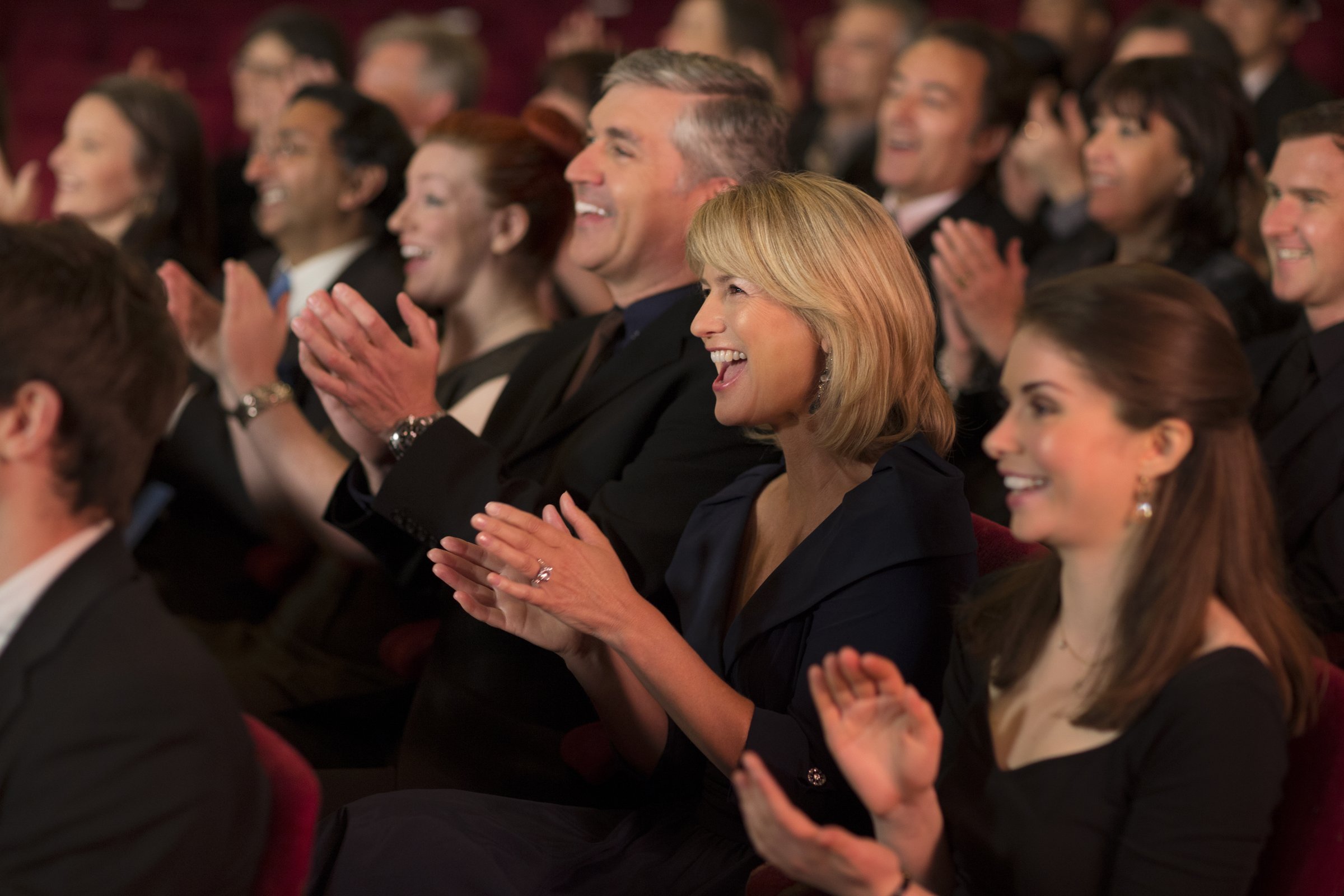 Audience in a theater clapping and smiling, including men and women dressed formally, seated closely in rows.