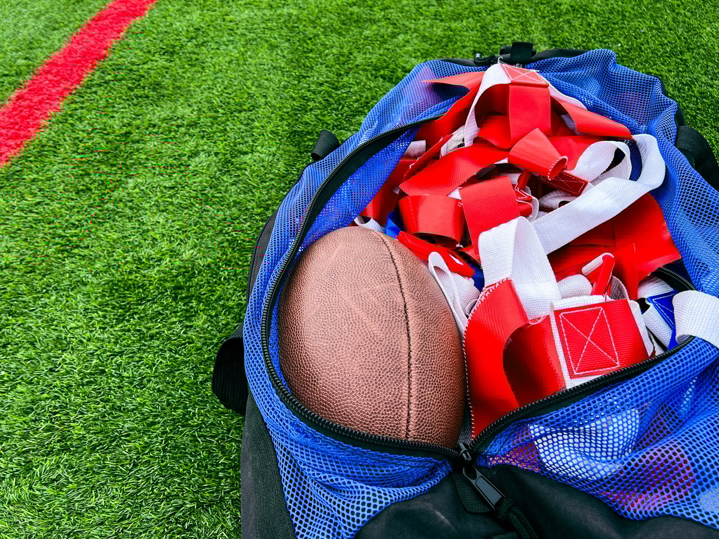 A large athletic duffel bag is filled with equipment for flag football play. Red and blue flags and a football fill the bag. Green turf football field background.